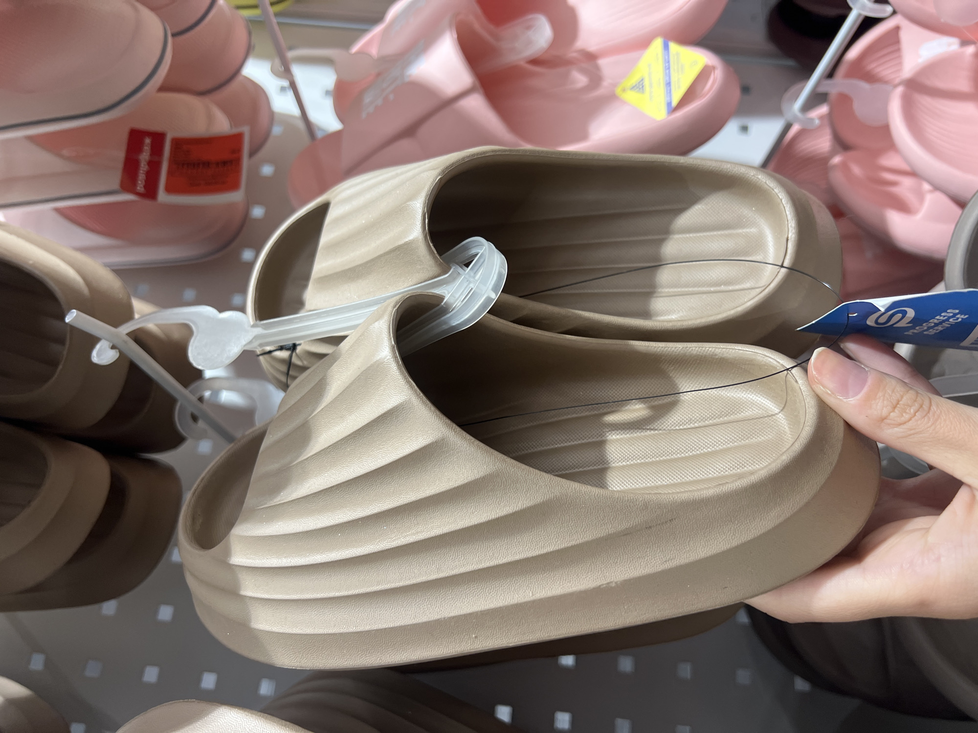 A light-skinned hand holds a pair of new, light beige (taupe) rubber or EVA foam slide sandals in a retail store setting in Kyiv, Ukraine. The sandals feature a textured, ribbed upper strap with a wave-like pattern and a similarly textured footbed. They are attached to a clear plastic hanger, and a blue tag with a white 'S' logo and "PROGRESS SERVICE" text hangs from a black string connected to the sandal. In the background, numerous other identical slide sandals in a slightly darker brown are visible hanging above. To the right, multiple pairs of pink versions of the same slide sandals are displayed, also on clear plastic hangers. One pink sandal has a blurry red price tag, and another shows a yellow tag with a blue triangular logo. The products are displayed against a white pegboard or slatwall with square perforations, suggesting a typical retail environment with bright, uniform lighting.
