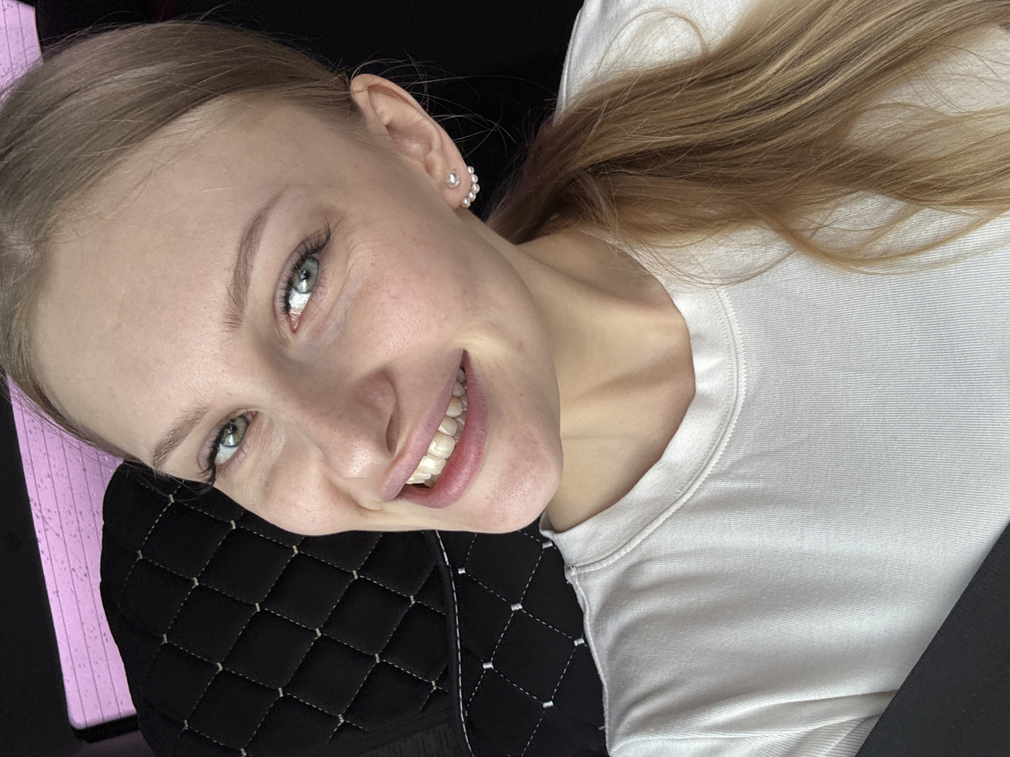 A blonde woman with light skin and green eyes smiles at the camera. She wears pearl earrings and a white t-shirt. She leans against a black quilted surface, possibly a car seat. The scene appears to be inside a vehicle. There is nothing to indicate that this photograph was taken in Perm, Russia.