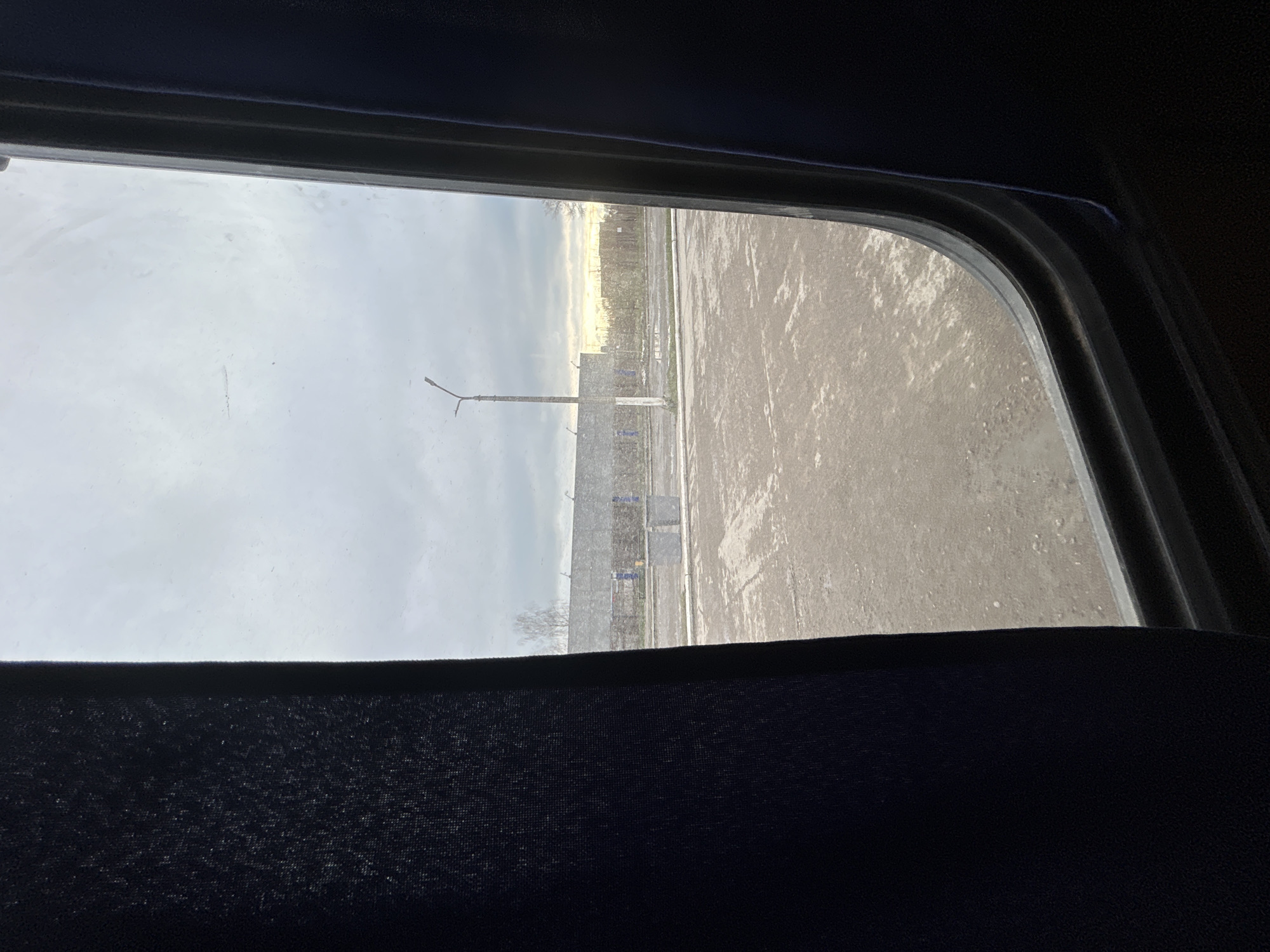 The image captures a view from inside a vehicle, looking out through a dark-framed window. The window's bottom edge is prominent, revealing a dark, textured fabric, likely an interior lining or curtain.
Outside the window, the scene depicts a semi-urban or industrial area under an overcast sky. The ground surface, which takes up a significant portion of the lower right, appears to be an asphalt or concrete road or parking lot. It has a dull, possibly wet or icy texture, suggesting cold weather. In the middle-left, a tall, slender lamppost or streetlight stands, featuring two horizontal light fixtures near its top. Behind the lamppost, a dark fence or barrier is visible, separating the foreground from a strip of bare trees or sparse vegetation.
On the distant horizon, a narrow band of bright, diffused light suggests either a low sun during sunrise or sunset, or sunlight breaking through a dense cloud cover. The sky above is uniformly light gray and hazy, indicative of an overcast day.
There are no people visible, and no specific activity or event is taking place beyond the static observation from the window. The overall atmosphere is quiet and subdued, influenced by the muted light and cool weather conditions. No visible text can be discerned. The bare trees and ground conditions are consistent with a cold climate, typical of locations like Chaikovsky, Russia, during winter or early spring.