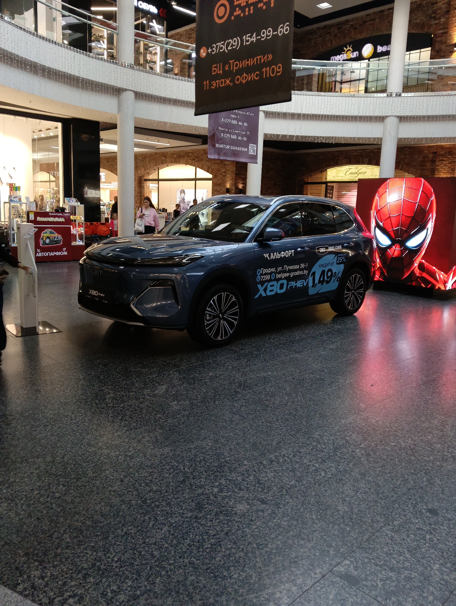 The image shows the interior of a shopping mall with a dark grey tiled floor. In the foreground, a dark blue Geely X80 PHEV is parked. The car has a sticker on its side advertising a loan rate of 1.49% from BELGEE. Another sticker on the car indicates "Grodno, ul. Luchkova 36-1, 7299 belgee-grodno.by". A large digital screen displaying a close-up of Spider-Man's face is positioned to the right of the car, casting a red glow on the floor. In the background, there are shops on multiple levels, with escalators and balconies visible. People are sparsely scattered throughout the mall. There is a black banner with orange text at the top of the image, which appears to be an advertisement.