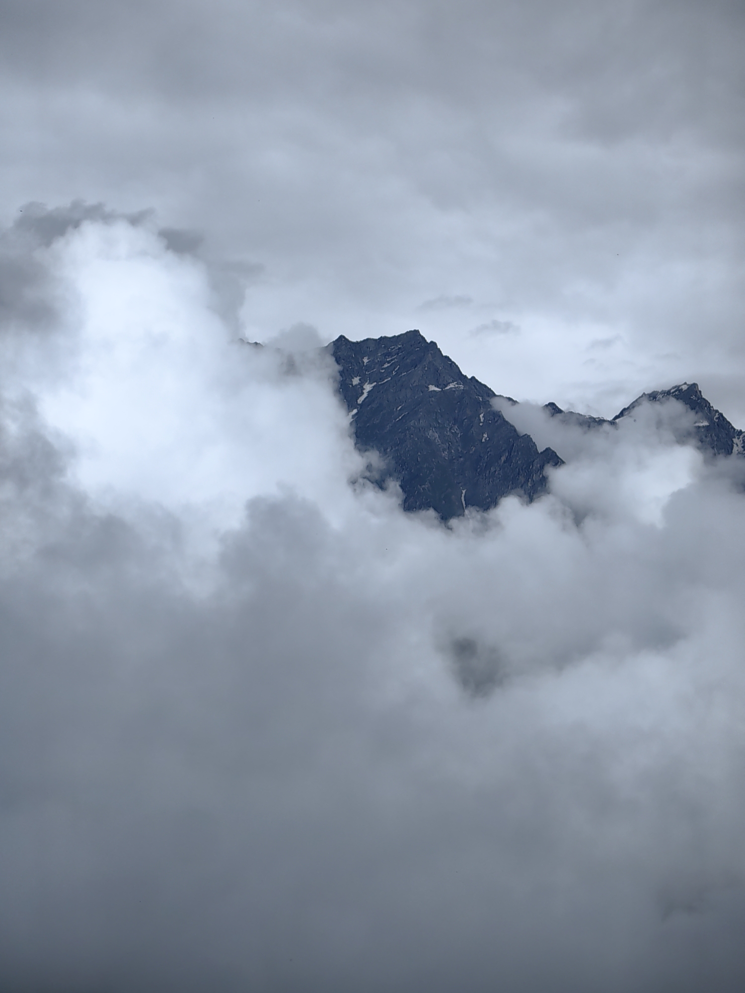 The image depicts a mountain peak shrouded in clouds. The mountain is dark and rocky, with patches of snow visible on its slopes. The clouds are thick and white, obscuring the lower portions of the mountain and creating a sense of depth and mystery. The sky above is overcast, with a muted gray color that suggests a cloudy or foggy day. The overall impression is one of a dramatic and majestic natural landscape.