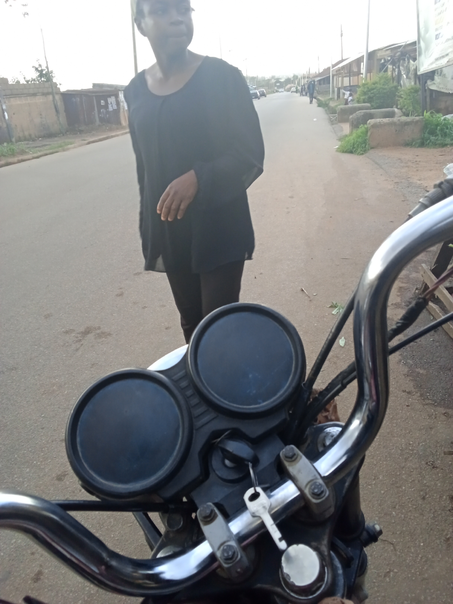 In Gura Top, Nigeria, a person in a black top stands on a road beside a parked motorcycle. The motorcycle's handlebars and instrument cluster are in the foreground, with two circular black gauges. The person is facing away from the motorcycle, looking towards the right side of the frame. The road is paved and appears to be in a moderately populated area, with buildings and vegetation visible along the sides. Several cars are parked or driving on the road in the background. The sky is overcast.