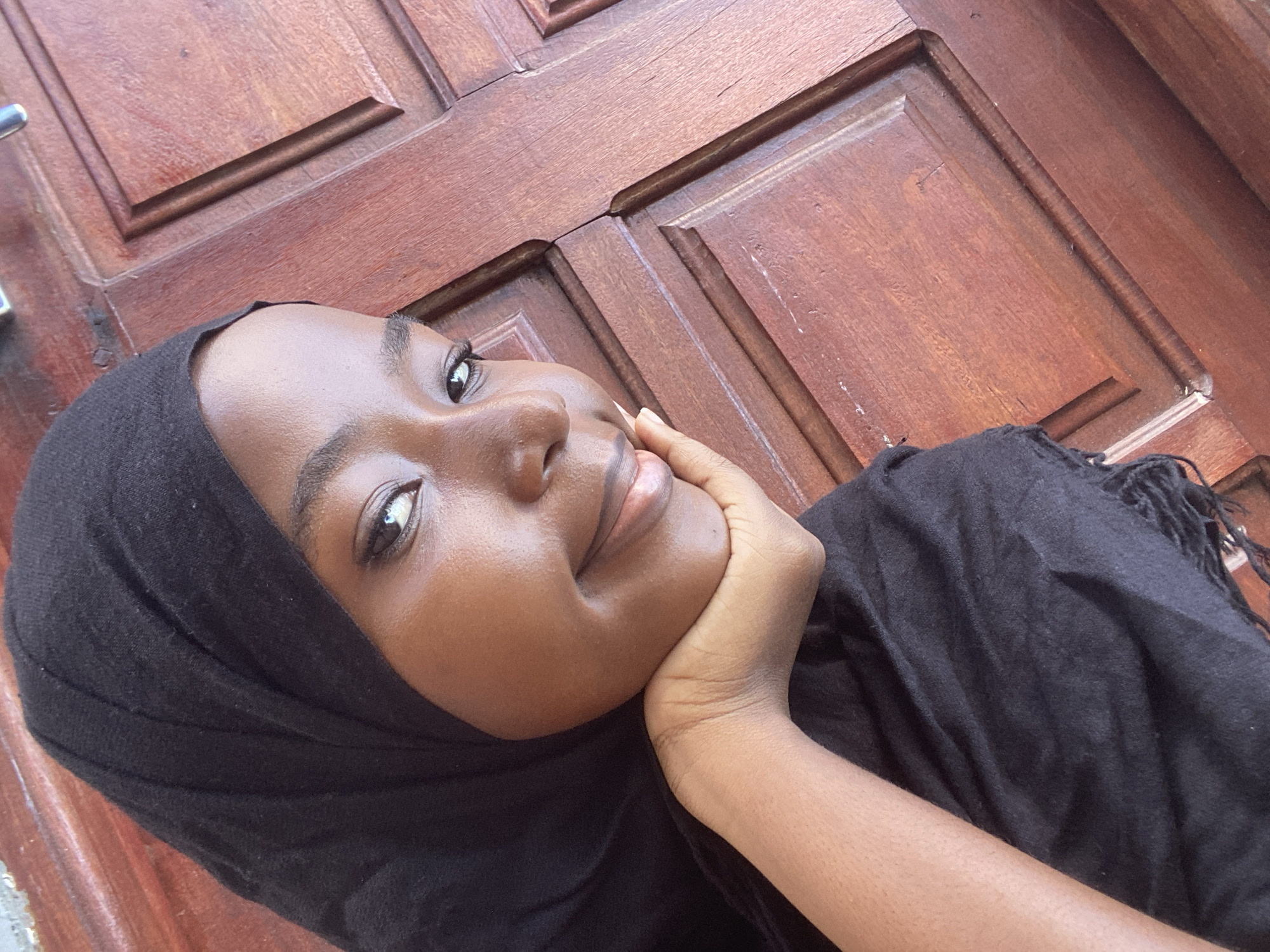 A close-up, angled portrait features a young individual, appearing female, with medium-dark skin. The person is positioned in the left foreground, looking directly at the camera with a subtle smile. Their left hand supports their chin, with the palm resting beneath and fingers curled against the cheek and jawline. They wear a black head covering, possibly a hijab, which drapes over their left shoulder, obscuring the rest of their body. The fabric of the head covering is plain black, with visible fringe at the bottom left. The background consists of a reddish-brown wooden door with multiple recessed panels, indicating an indoor or sheltered setting. A vertical seam or joint is visible in the wood paneling to the right of the individual's face. The specific location within Dar es-Salaam, Tanzania, cannot be determined from the visual information provided.