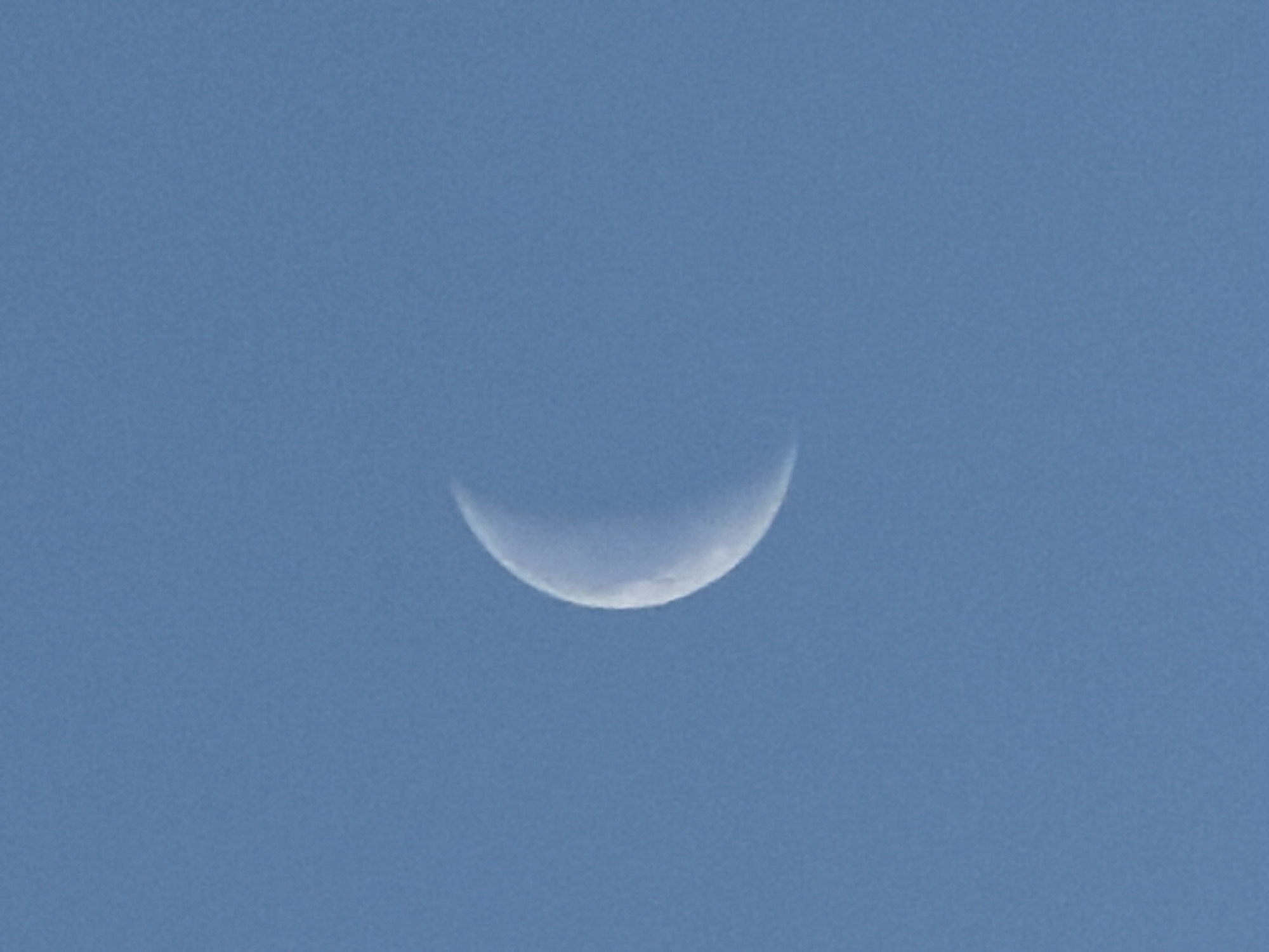 The image shows a crescent moon in a blue sky. The moon is a thin sliver, illuminated on its lower edge. The rest of the moon is faintly visible due to earthshine. The sky is a uniform, clear blue. The location is not provided.