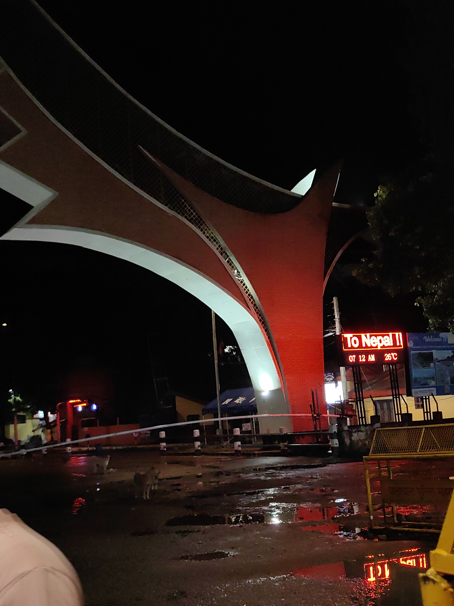 At night in Belahiya, Nepal, a modern, arching structure made of reddish-brown brick and white material spans across a wet, reflective street. A digital sign on a pole illuminates the words "To Nepal II" in red, along with the time "07:12 AM" and the temperature "26°C". The street surface shows numerous puddles reflecting the red light from the sign and other unseen sources. In the background, a red truck with blue lights is visible, along with a yellow metal barrier. Two dogs stand on the wet street, one looking towards the camera. Trees with foliage are present on the right side of the frame, and a dark sky suggests it is nighttime. A portion of a person's shoulder, clad in a light-colored garment, is visible in the lower left corner of the image.