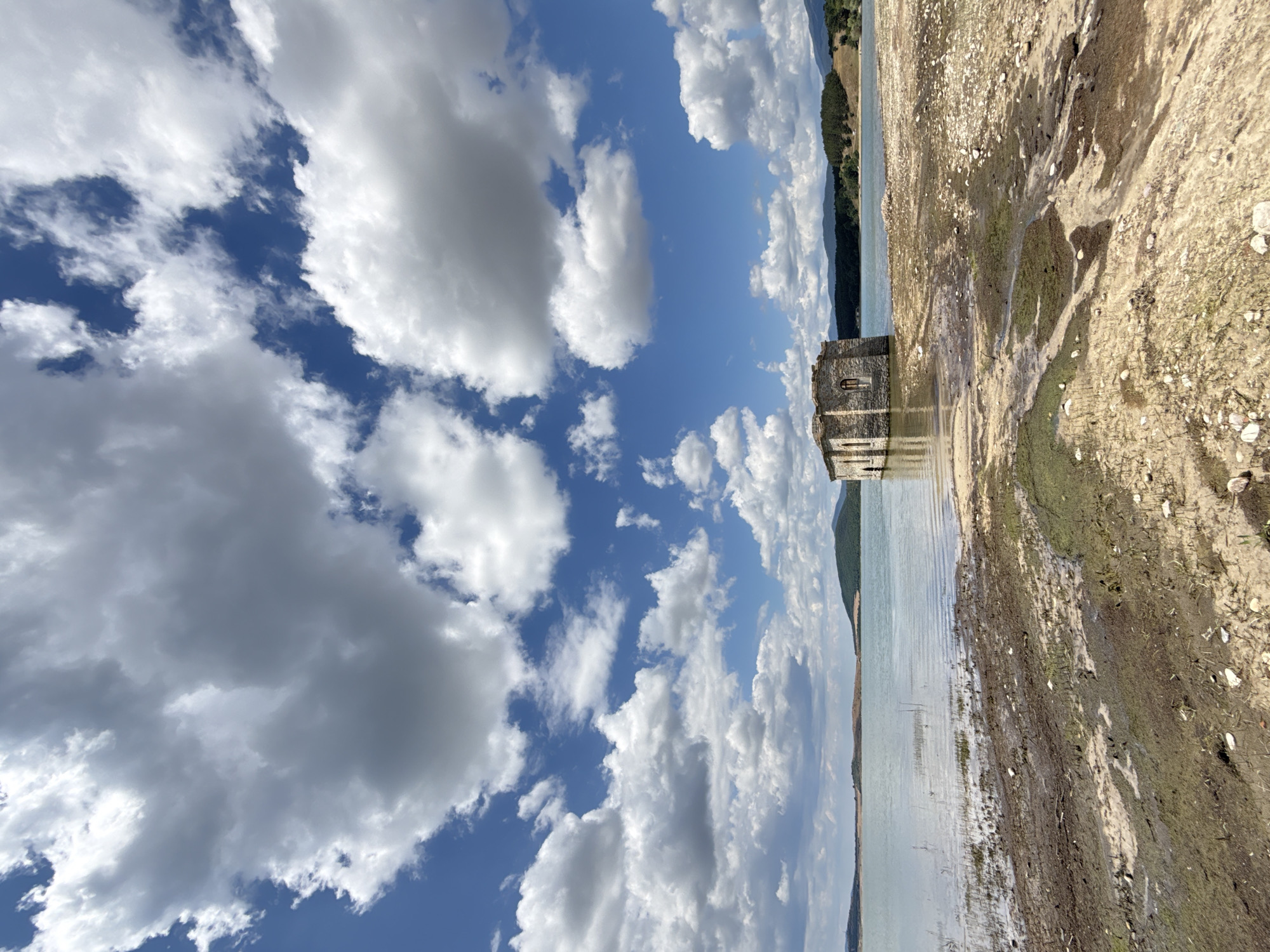 The image depicts the St. Ivan Rilski Chapel, submerged in the Zhrebchevo Reservoir near Panicherevo, Bulgaria. The stone structure, partially visible above the water's surface, exhibits weathered stone walls and arched windows. The surrounding environment is a body of water with visible ripples, extending towards a distant shoreline characterized by rolling hills and a tree line. The foreground reveals a dry, muddy bank littered with stones and sparse vegetation, indicating a low water level. The sky above is a vibrant blue, filled with large, fluffy white clouds, some casting shadows on the landscape. The overall scene evokes a sense of solitude and abandonment.