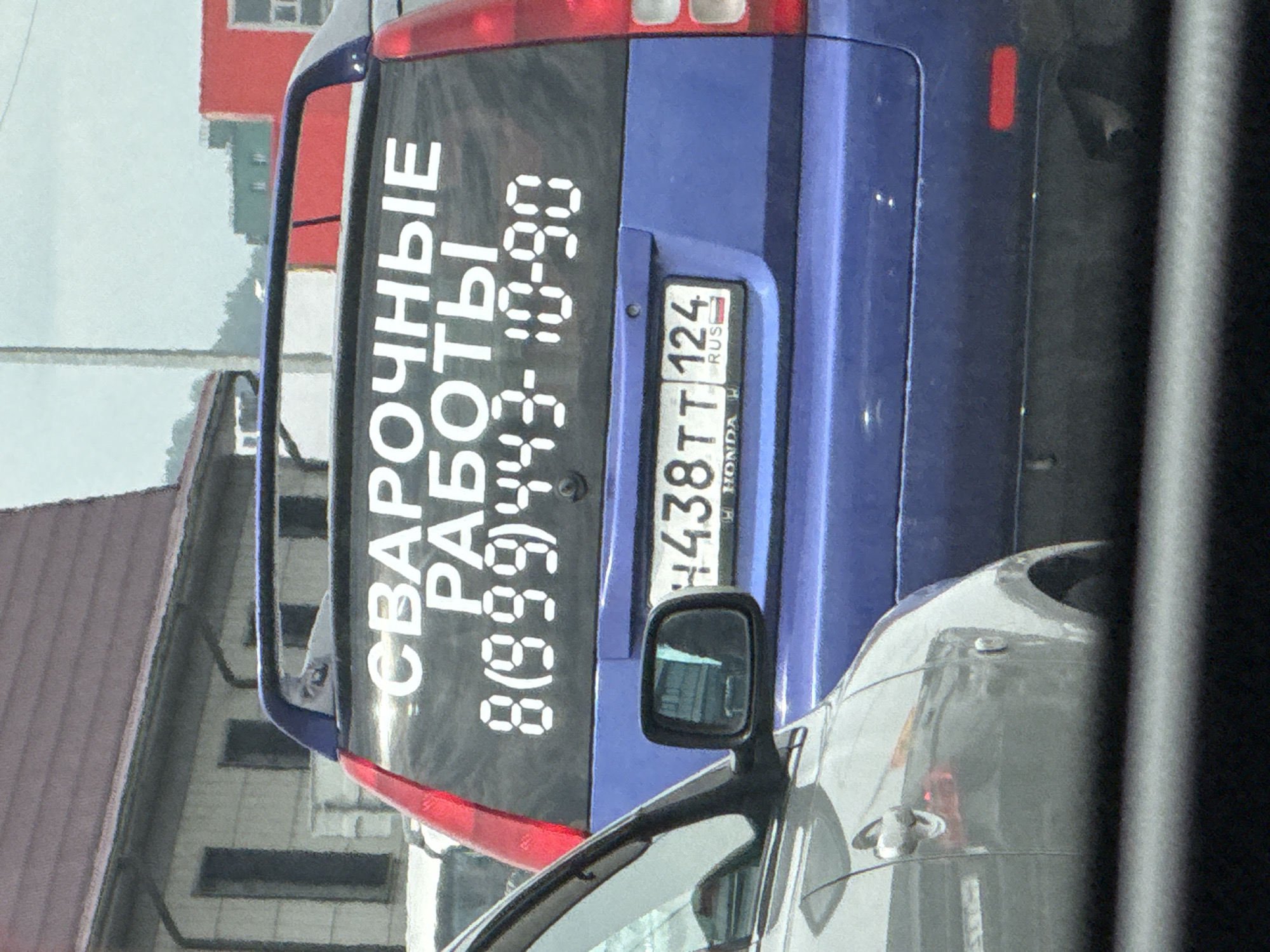 The image shows the rear of a blue car parked on a street. On the rear window, there is a black sticker with white text that reads "СВАРОЧНЫЕ РАБОТЫ 8(999)443-10-90". This translates to "Welding Services 8(999)443-10-90". A license plate is also visible on the rear of the car, reading "438TT 124 RUS". The scene appears to be in a residential area, with a building with a red roof and white walls visible in the background. The sky is overcast, suggesting a cloudy day. The overall impression is of a utilitarian vehicle advertising a service. There are no people visible in the image.