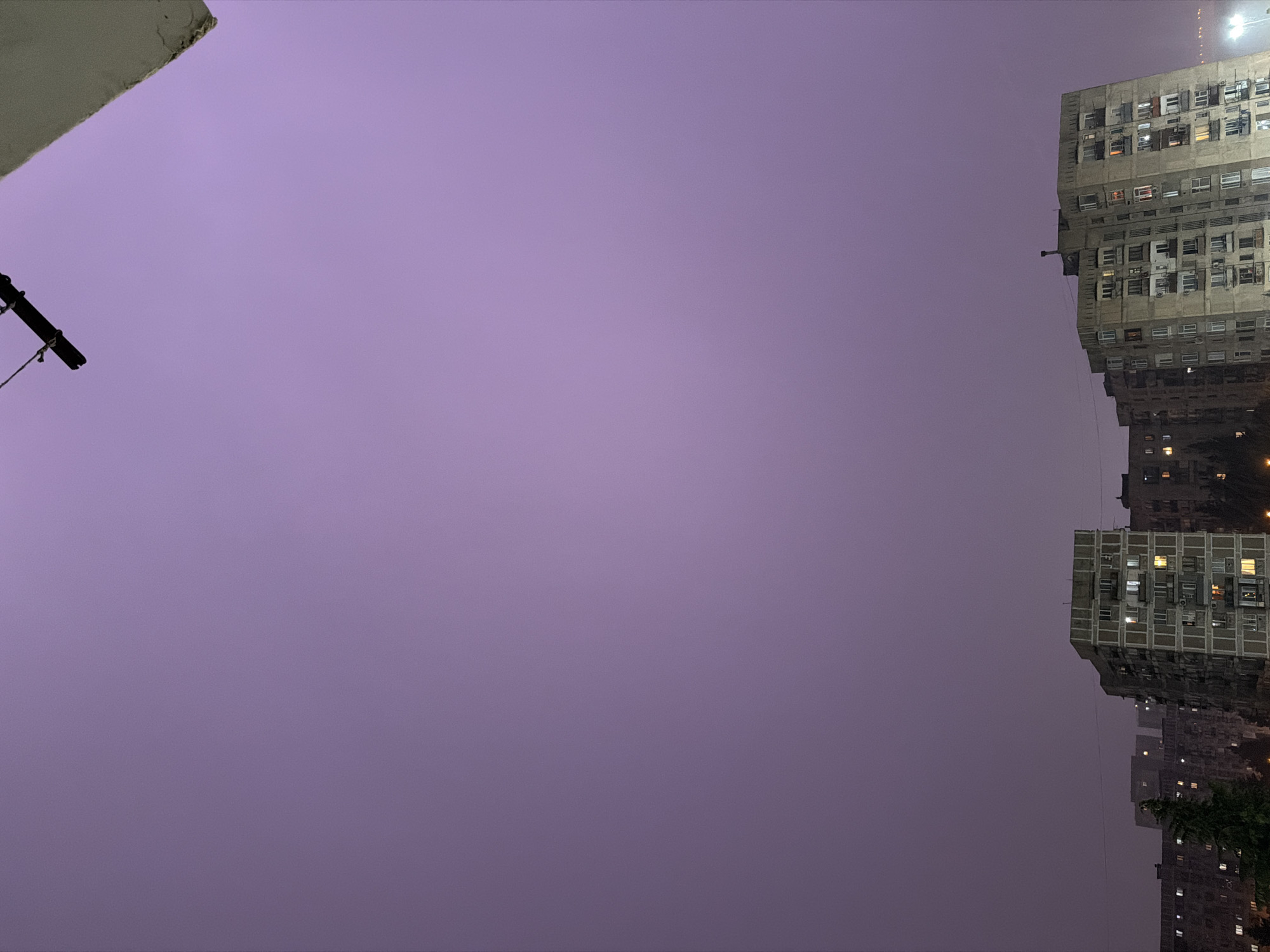 The image is a low-angle shot of buildings in Tbilisi, Georgia, taken at night. The sky is a purple-grey hue. On the right side of the frame are several high-rise apartment buildings constructed of grey concrete. Windows are visible in the buildings, some illuminated with light. In the upper left corner, there is a partial view of a white roof and a black pole extending into the sky. The buildings appear to be located within a densely populated area, indicative of an urban environment.