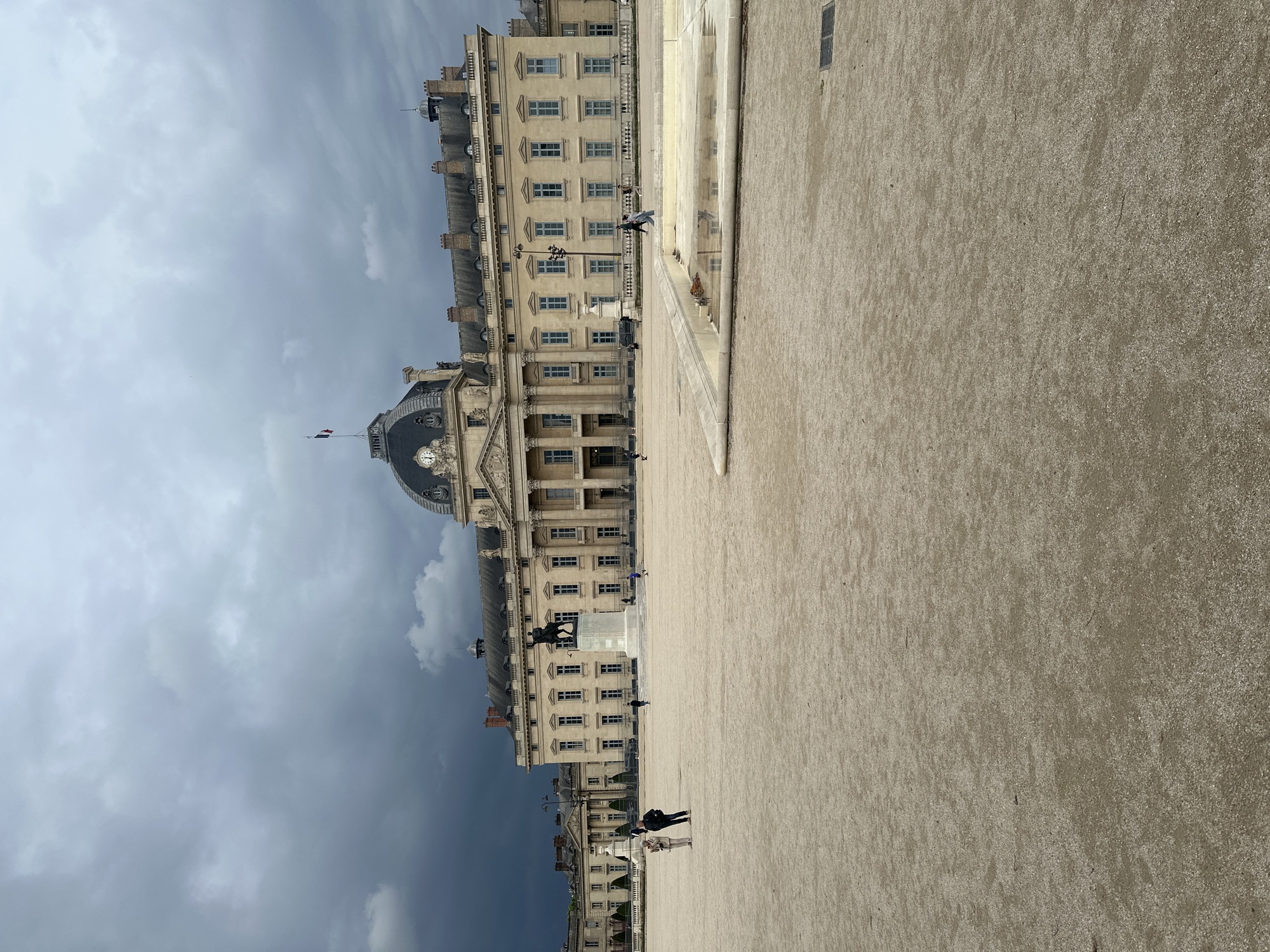 The image shows the Les Invalides complex in Paris, France. The scene is dominated by a large, light-colored building with many windows. The building has a prominent dome. The sky above is overcast and grey. The foreground is a wide, open space paved with gravel. Several people are walking in the open space.