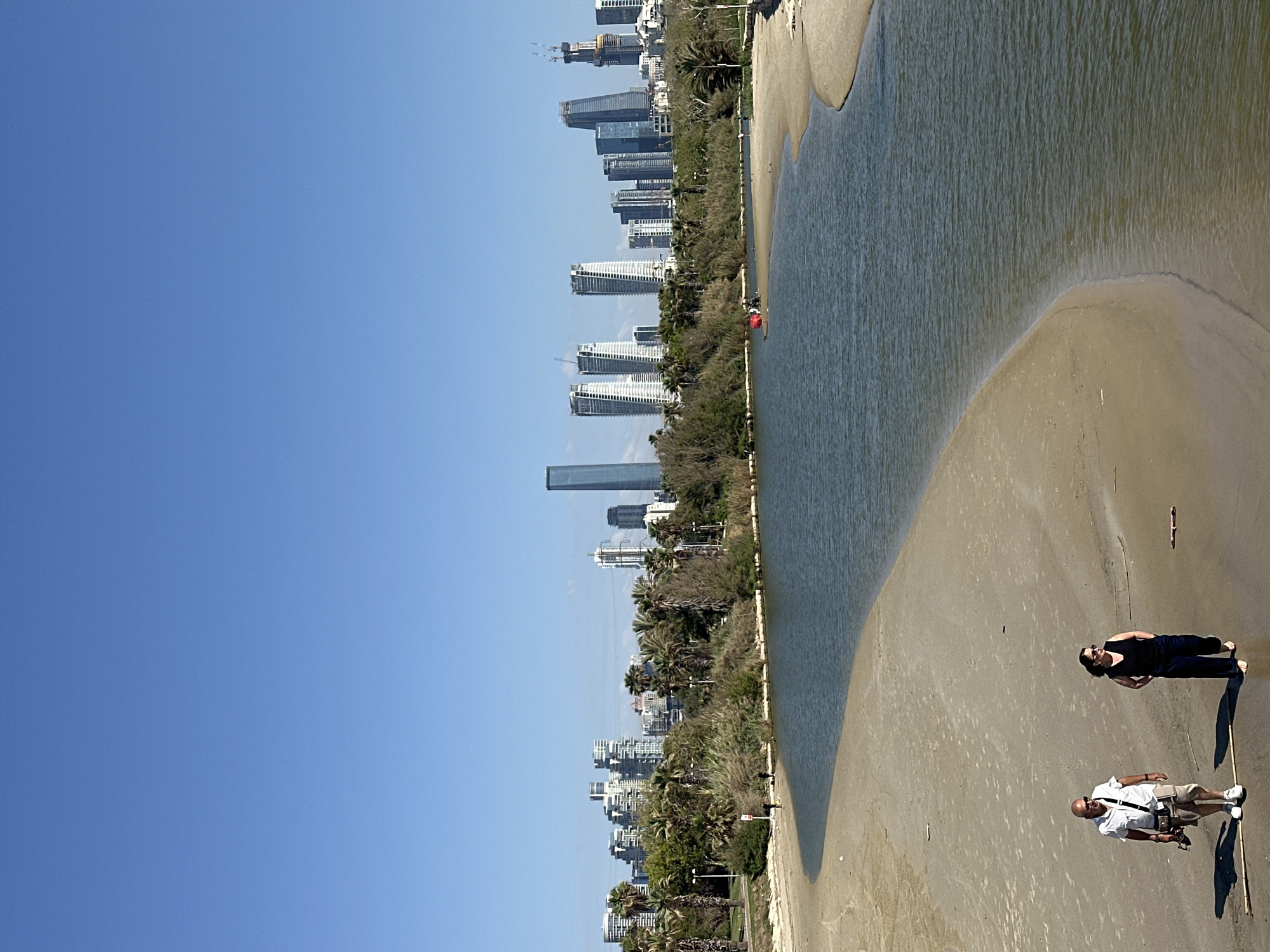 The image shows the Yarkon River as it flows into the Mediterranean Sea in Tel Aviv, Israel. The scene is divided vertically between a clear, blue sky on the left and the river, beach, and cityscape on the right. The city's skyline, featuring numerous modern high-rise buildings, runs along the left side of the river. The sand is wet near the water, becoming drier towards the bottom of the image. Two people are visible on the beach at the bottom right: one is lying down, and the other appears to be standing and taking a picture with a camera. A shadow of a structure, likely a bridge or viewing platform, falls across the river and beach. Trees line the riverbank, separating the city from the river and beach.