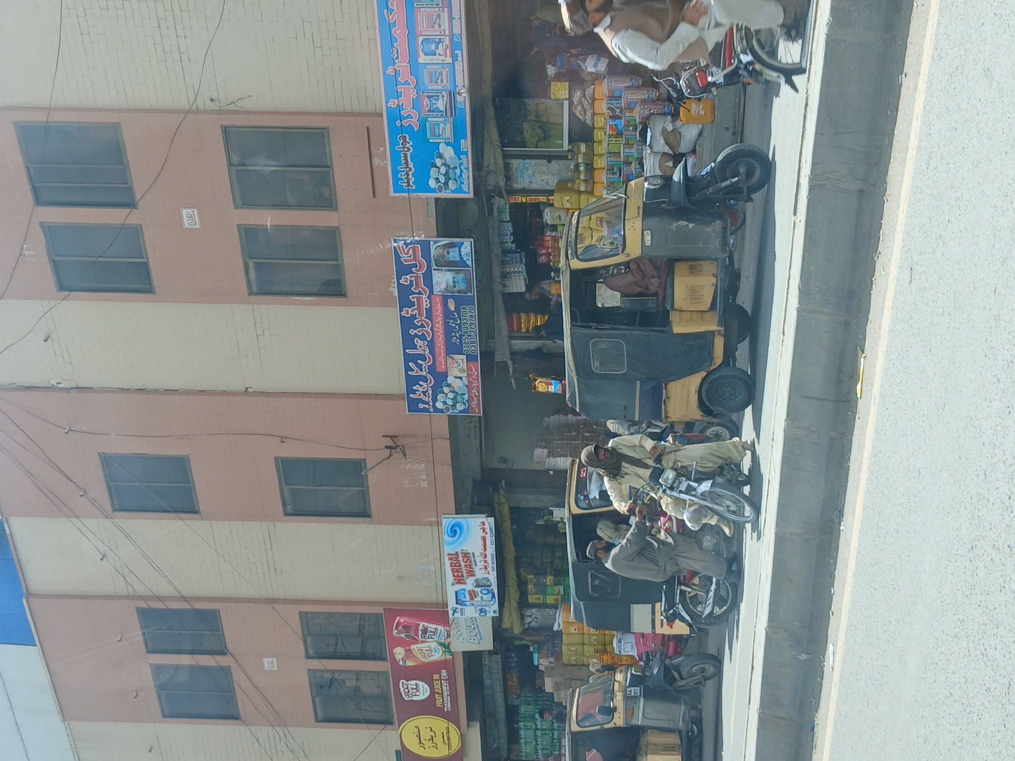 This image captures a bustling street scene in Quetta City Tehsil, Pakistan, bathed in bright daylight. The scene features multiple men, largely dressed in traditional shalwar kameez, navigating or idling near vehicles. Several auto-rickshaws, distinctive yellow and black three-wheelers, are prominent, along with numerous motorcycles.
The activity depicted is typical daily urban life in a commercial district, with people and vehicles moving along the street. Shops at the ground level of multi-story buildings display various goods, creating a vibrant retail environment. The clear, sunny weather casts distinct shadows across the street and buildings.
Notable details include the characteristic South Asian architecture, the style of the vehicles, and the attire of the people, all consistent with the Pakistani context. Overhead, numerous power lines crisscross the view.
Visible text on various signs and advertisements, predominantly in Urdu, includes "منصور ٹریدرز" (Mansoor Traders), "FRUIT JUICE IN TRANSPARENT CAN", "حکمت ٹریڈرز" (Hikmat Traders), and "گل ٹریڈرز" (Gul Traders). A sign also advertises "HERBAL WASH" and mentions "حاجی من عصمت الله تر يفرز" (Haji Min Ismatullah Tariq Nawaz/Tarif Nawaz) with phone numbers "0305-8093708" and a partial "03". The word "BOUND" is also visible on one of the signs.