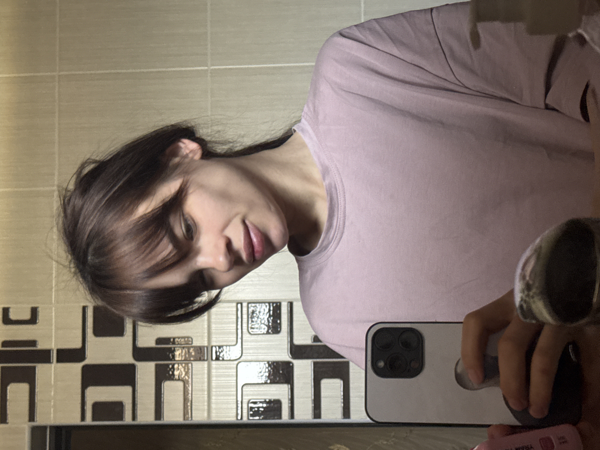 A woman is taking a selfie in a bathroom. She has brown hair with bangs, and she is wearing a light purple shirt. She is holding a white smartphone with three camera lenses. The background is a bathroom wall with beige and brown patterned tiles. The bathroom is likely located in an apartment building in Rostov-on-Don, Russia. There is a bottle of Nivea cream.