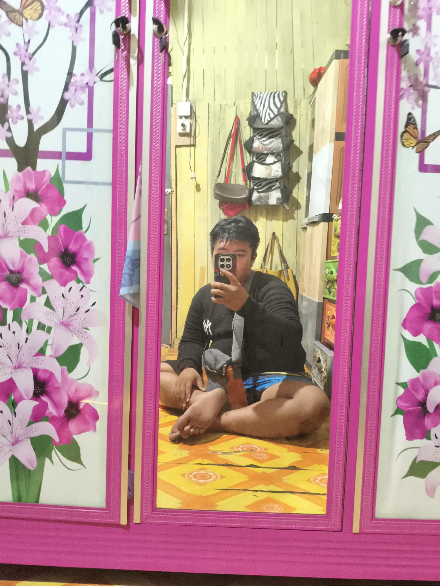 A young adult male, with short dark hair, is sitting cross-legged on a yellow and orange patterned floor, taking a selfie in a mirrored wardrobe. He is wearing a black long-sleeved shirt with a white logo on the left chest, a grey crossbody bag, and blue patterned shorts. The wardrobe has pink frames and decorative panels featuring pink and white flowers and a butterfly. The background visible in the reflection includes a wall with horizontal wooden planks, several items hanging on the wall, including a zebra-print hanging organizer, a brown handbag, and a yellow tote bag. To the right of the wardrobe, there are shelves with colorful objects. The scene appears to be an indoor residential space, likely a bedroom. There is no discernible information indicating the specific location within the city of Samarinda, Indonesia.