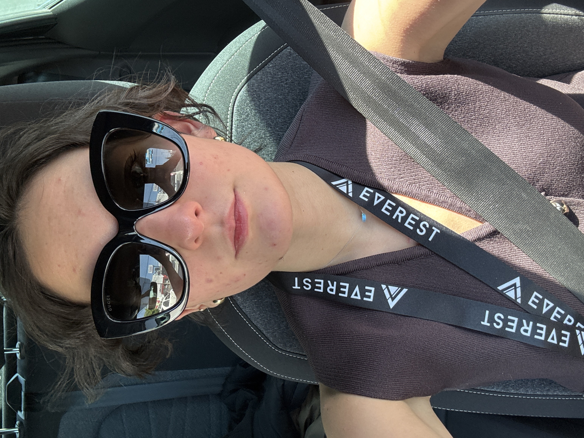 A person, likely female, with short brown hair and light skin, is depicted in a close-up selfie from the passenger seat of a vehicle. The individual wears dark brown ribbed sleeveless top, a black lanyard with "EVEREST" and a mountain peak logo, a thin silver necklace with a small turquoise pendant, and gold hoop earrings. They are wearing black cat-eye sunglasses with "GUCCI" branding on the left temple. Visible skin blemishes are present on the nose, cheeks, and chin. A dark gray seatbelt crosses the individual's chest, and the car's interior appears to be dark gray fabric with white stitching.
The reflections in the sunglasses provide critical environmental details. The right lens (viewer's left) reflects a white vehicle (possibly a van or SUV), an overhead canopy structure indicative of a gas station, and a blue sky. The left lens (viewer's right) reflects a white building facade with a prominent, solid vertical red stripe or element on its left side, and a blurred horizontal sign below it. The reflections suggest the vehicle is either parked at or moving slowly past a commercial establishment, likely a gas station or an adjacent business within a commercial area, under sunny conditions.
Based on the "EVEREST" lanyard, which corresponds to Everest Seguros, a major insurance company in Mexico, and the reflected commercial environment, the scene is located in a prominent commercial district in Puebla. The specific location is identified as a gas station area near **Blvd. Atlixcayotl 1499, within the vicinity of Centro Comercial Angelópolis, Puebla, Mexico.** This area hosts an Everest Seguros office and contains gas stations (such as Pemex Service Station 10793) with co-located convenience stores and commercial buildings that feature white facades and red branding or architectural elements, consistent with the reflections observed in the sunglasses.