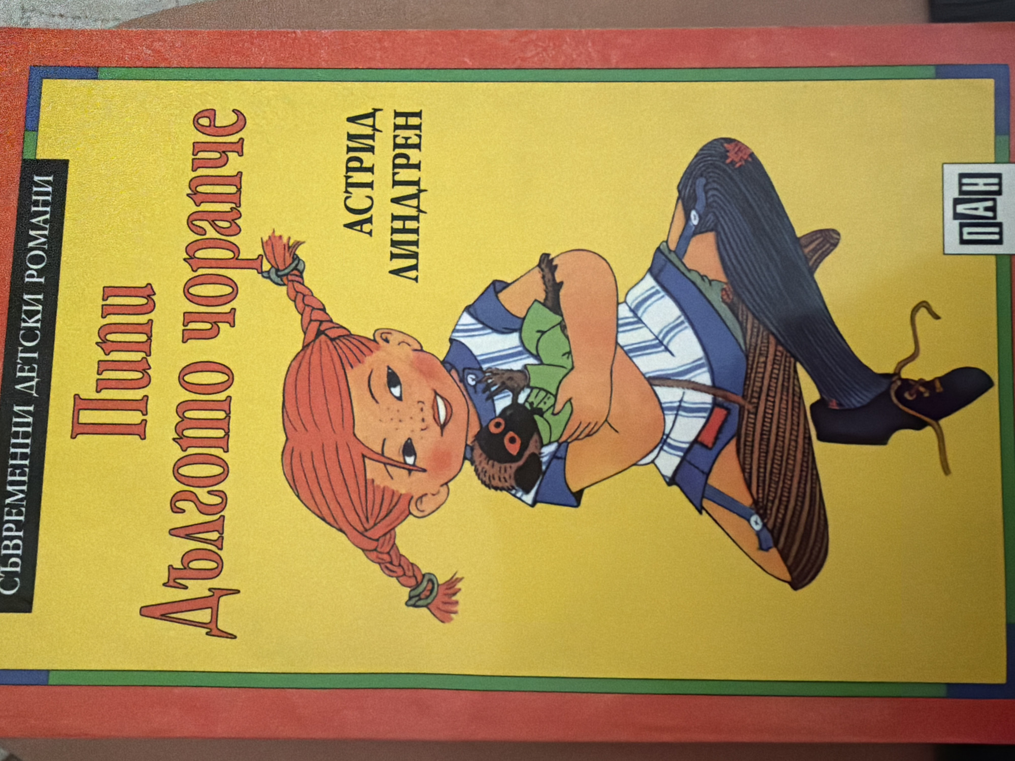 The image is a close-up of the cover of a book titled "Pippi Longstocking" in Bulgarian. The title is written in large, stylized red font. "Pippi" is at the top, followed by "Dalgomto chorapche" (meaning "long stocking") below it. At the very top, in smaller black text, it says "Sâvremenni detski romani" (meaning "Modern children's novels"). To the right of the illustration of Pippi, the author's name "ASTRID LINDGREN" is written in black capital letters. The illustration depicts a cartoon character, Pippi, with red braided hair, freckles, and a blue and white striped dress. She is holding a small monkey. The background is bright yellow. In the bottom right corner, there is a logo with the letters "ПАН" in white on a black background within a white rectangle.
This image is a book cover and therefore does not depict a specific location in the city of Pazardzhik, Bulgaria, or any real-world scene.