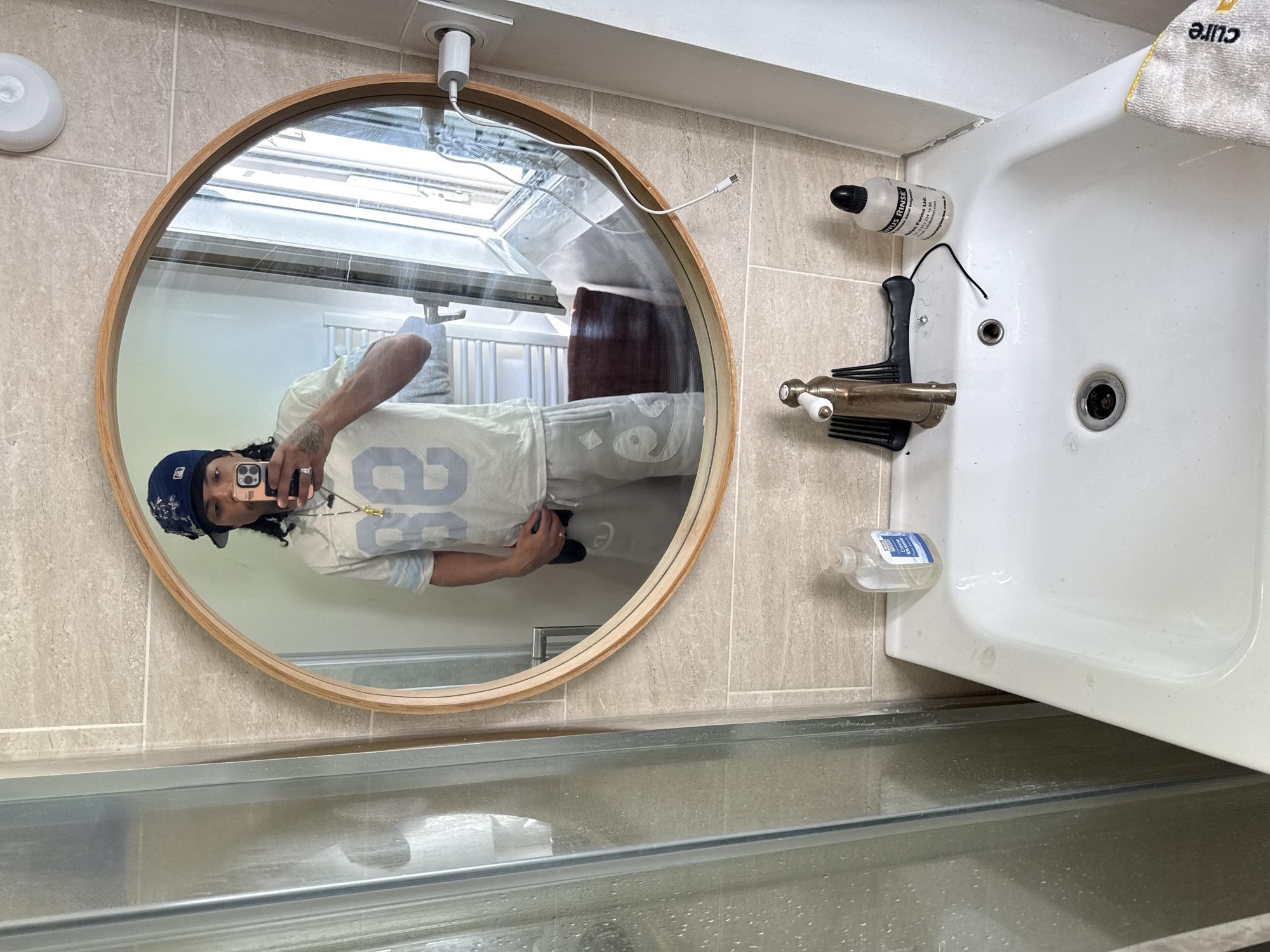 A full-body mirror selfie taken in a bathroom shows a person with dark hair, wearing a blue baseball cap, a light-colored jersey with the number "88" in blue, and light-colored shorts. The person is holding a phone in their right hand, taking the photo. The bathroom features light-colored tiled walls, a white sink with a faucet, a bottle of soap, a comb, and a bottle of nasal rinse. A shower enclosure is visible on the left side of the image, and a window is reflected in the mirror. The scene appears to be a personal bathroom in Massy, France.