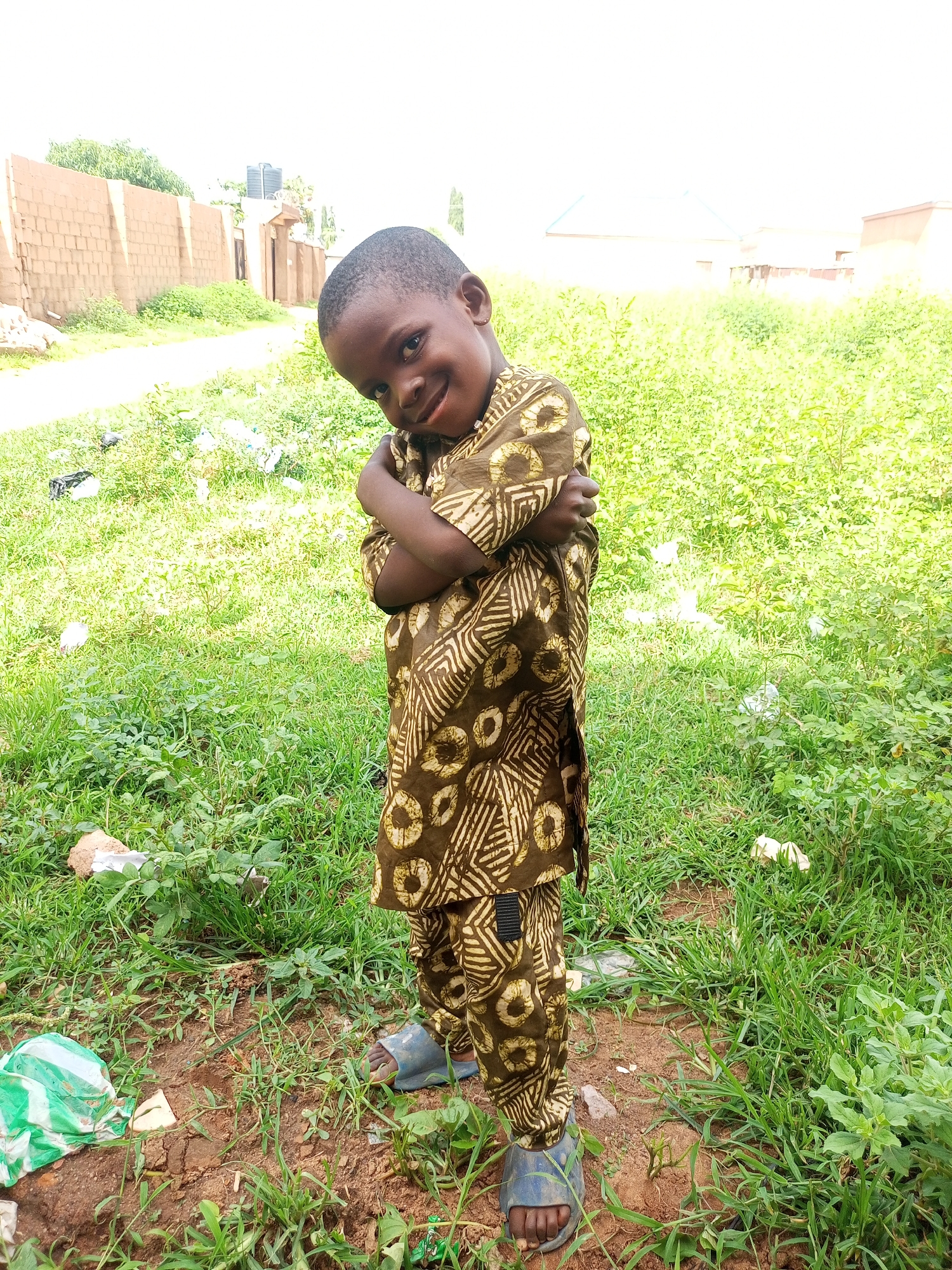 A young boy stands in an outdoor setting with his arms crossed and a slight smile. He is wearing a patterned brown and tan outfit, consisting of a shirt and pants. The boy is positioned in the foreground, with a background of overgrown green grass and a dirt path. In the distance, a wall constructed of light brown bricks is visible, with some vegetation growing near it. Further in the background, a building with a light blue roof can be seen. The ground around the boy is a mix of dirt and grass, with some scattered litter, including plastic bags. The lighting suggests it is daytime, with bright natural light. The location is identified as the city of Tempure, Nigeria.