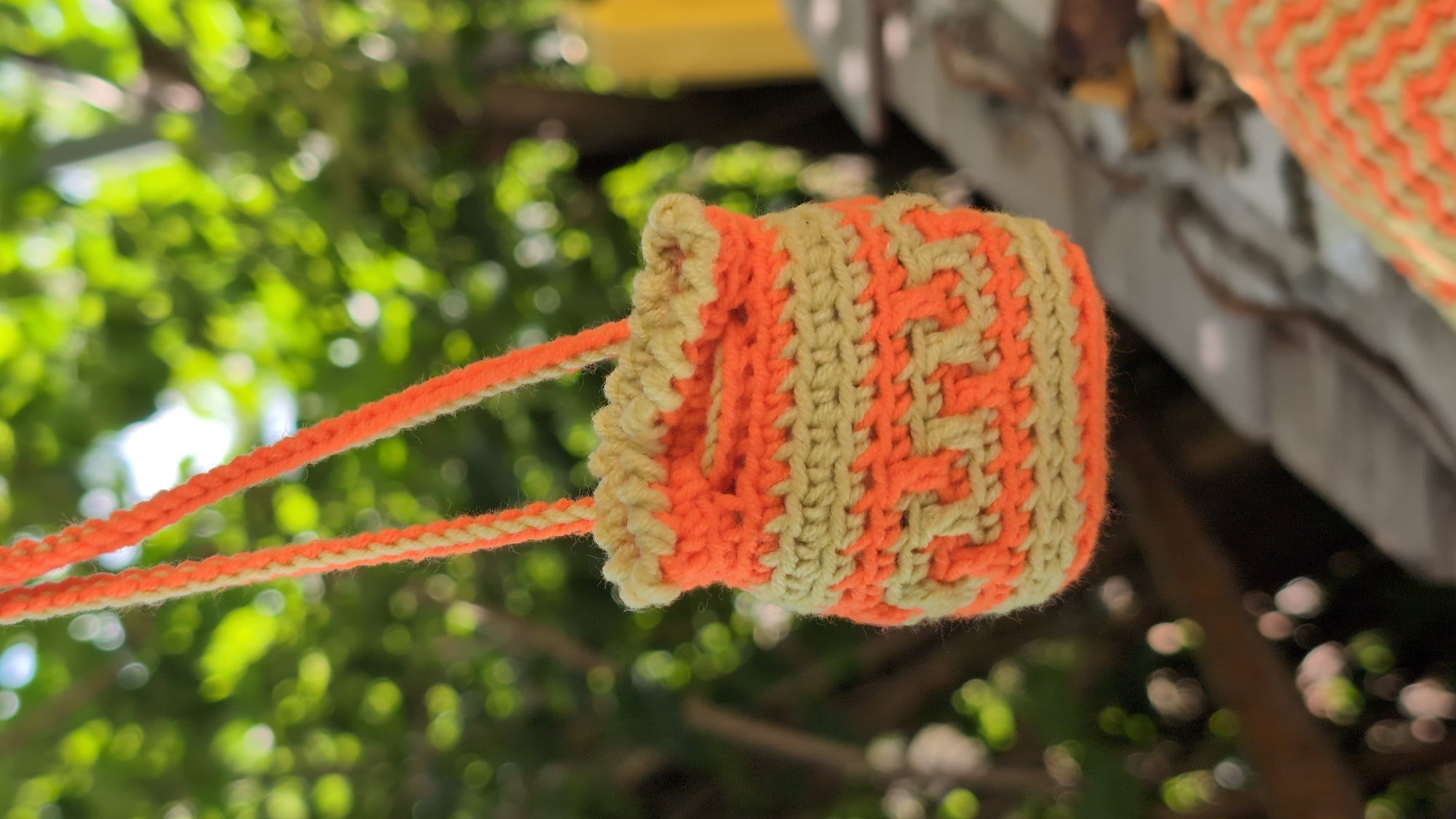 An orange and tan crocheted bag with a patterned design hangs from two crocheted straps. The bag is small and cylindrical, with a ruffled top edge. The background is blurred and consists of green foliage, suggesting an outdoor setting. The image does not provide any information about the city of Kostiantynivka, Ukraine, or any specific location within it.