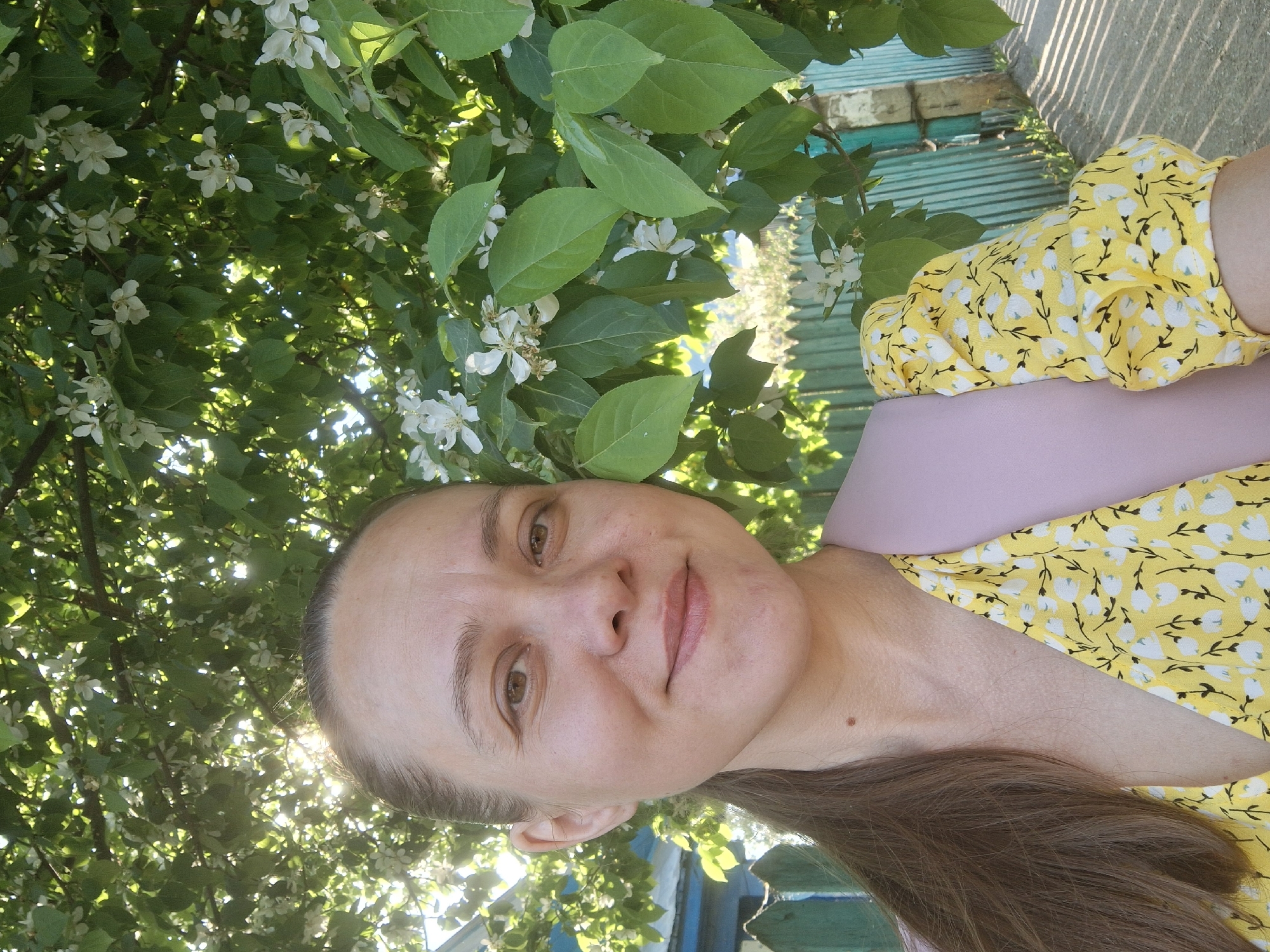 A middle-aged woman is posing for a selfie outdoors, possibly in Chaikovsky, Russia. She is wearing a yellow floral print dress with puffy sleeves and a pink top underneath. Her hair is brown and pulled back. She is standing beneath flowering trees, with leaves and white blossoms visible above and around her. In the background, there is a blue and green fence and a hint of sunlight filtering through the trees.