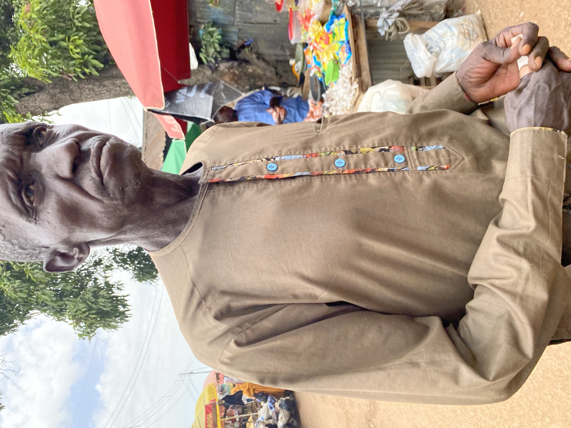 This media file shows a portrait of an elderly Black man in Bukuru, Nigeria. He is wearing a brown shirt with colorful, patterned fabric accents and blue buttons along the placket. His hands are clasped in front of him. The background appears to be a busy outdoor market scene, with stalls, colorful items, and other people visible. There are also trees and a cloudy sky. The lighting suggests it is daytime. The man's expression is serious, with his mouth slightly open. The overall scene depicts a moment in everyday life in a Nigerian marketplace. One stall in the background has visible text that appears to say "MAGGI".