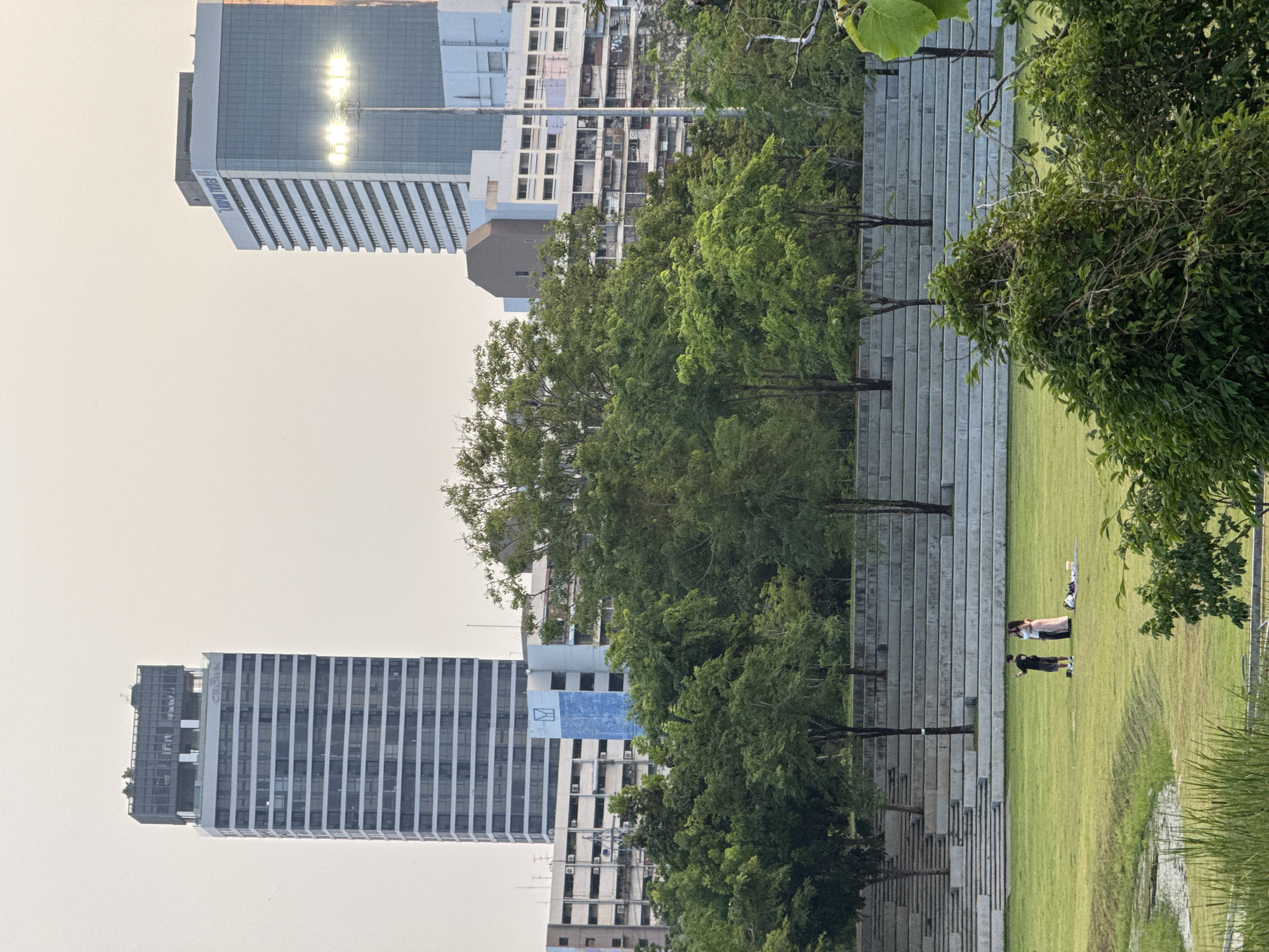The image depicts a park scene in Bangkok, Thailand. Two tall, modern buildings stand in the background; one has a reflective glass facade. Between the buildings and the park is a line of trees. A concrete tiered structure descends into a grassy area where two individuals are present. One person appears to be taking a photograph of the other. The scene is bathed in the soft light of either early morning or late afternoon.
