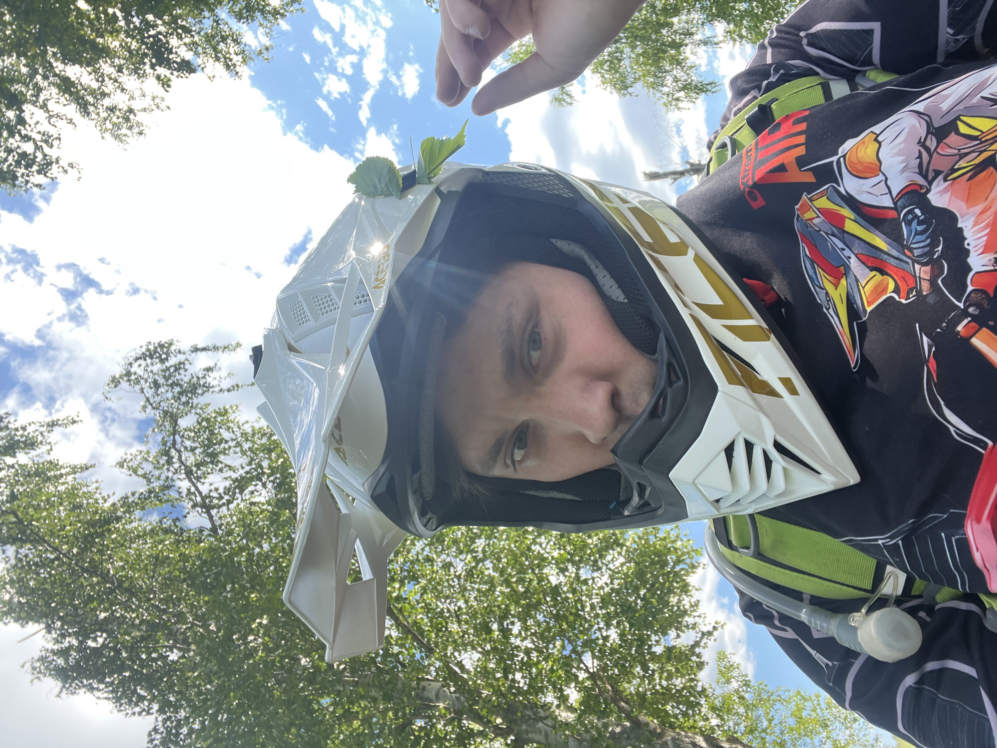 This low-angle, close-up outdoor portrait, taken in Новочувашский, Russia, captures an individual from the chest up, wearing a white and gold motocross helmet (branded "Acerbis") and a black long-sleeved jersey. The subject, likely male, looks directly into the camera; his face is partially obscured by the helmet, revealing his eyes, nose, and mouth, with dark hair visible underneath. A small green leaf is visibly tucked into the right side of the helmet, near the visor. A green-strapped hydration pack is visible over the left shoulder, with a clear plastic drinking tube and bite valve clipped to the front strap. The black jersey features a colorful graphic of a dirt bike rider and the text "Xtreme AIR" in red and white on the upper left chest. The background consists of a bright blue sky with scattered white cumulus clouds, framed by lush green foliage of deciduous trees, consistent with birch, particularly in the upper left. Strong sunlight illuminates the scene, casting highlights on the helmet and leaves. A hand, likely the subject's right, is partially visible in the upper right quadrant of the image, with fingers extended.