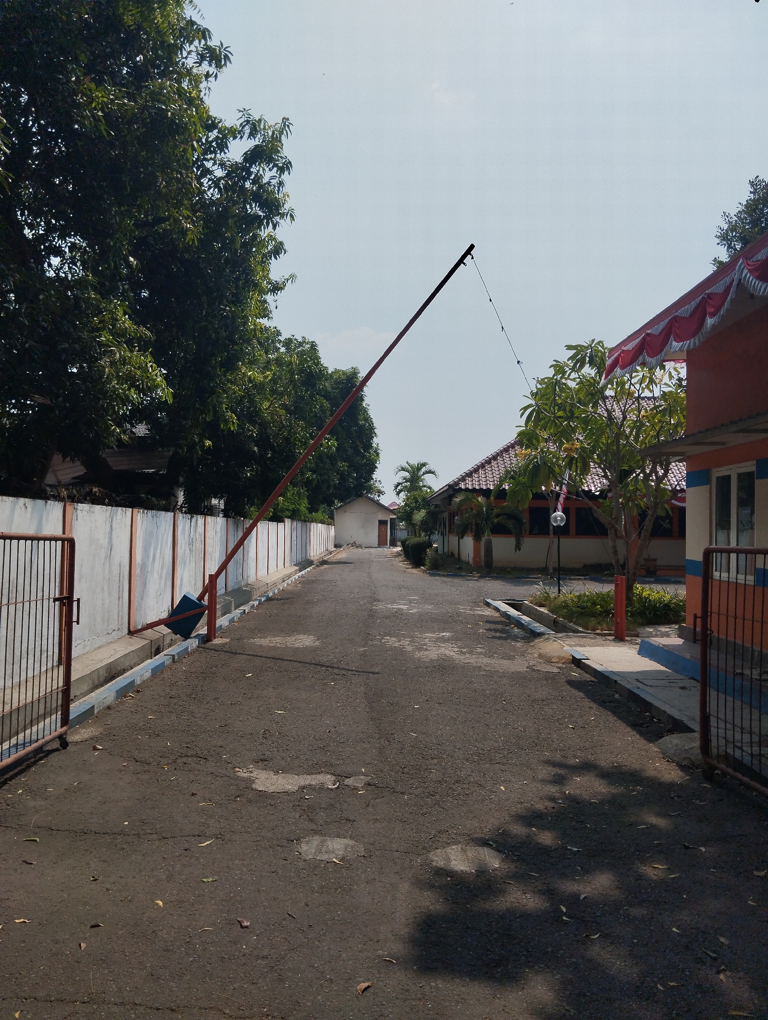 The image depicts an exterior view of a road leading into a complex with buildings and trees. On the left side of the road, there is a metal fence and a concrete wall with an orange trim. A red boom barrier is extended across the road, propped up by a blue object. Further down the road, on the right, is an orange and white building with a red and white decorative trim along its roofline. Green trees are visible throughout the scene, with some casting shadows on the asphalt road. The sky is clear and blue. This scene appears to be located in the city of Madiun, Indonesia, based on the architectural style and vegetation.