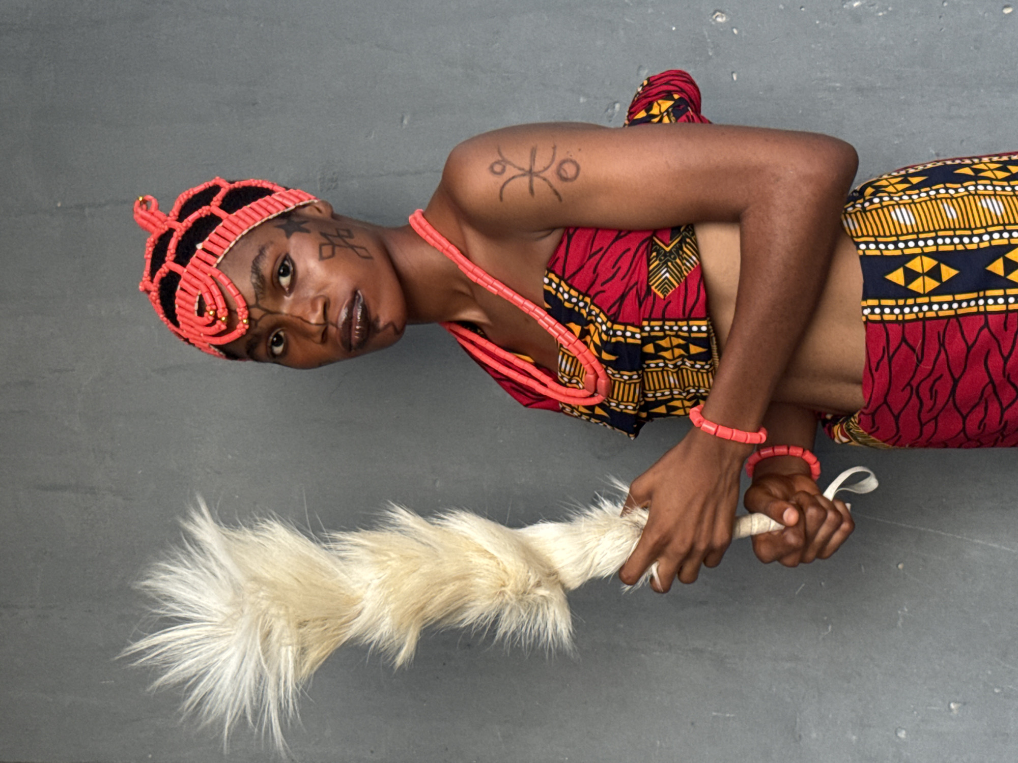 A medium shot shows a person wearing traditional Nigerian clothing and accessories lying on a gray surface. The person is positioned at a slight angle, looking directly at the camera.
The person is adorned with a beaded headdress in red and black, as well as facial markings. A red beaded necklace and two red beaded bracelets adorn their neck and wrists. A tattoo is visible on their upper arm. The person is wearing a red, yellow, and black patterned fabric wrapped around their waist. They are holding a white, feathery object in their hands.
The gray surface on which the person is lying appears to be a solid, neutral background. There is no other information about the environment to indicate a location in Badagry, Nigeria.