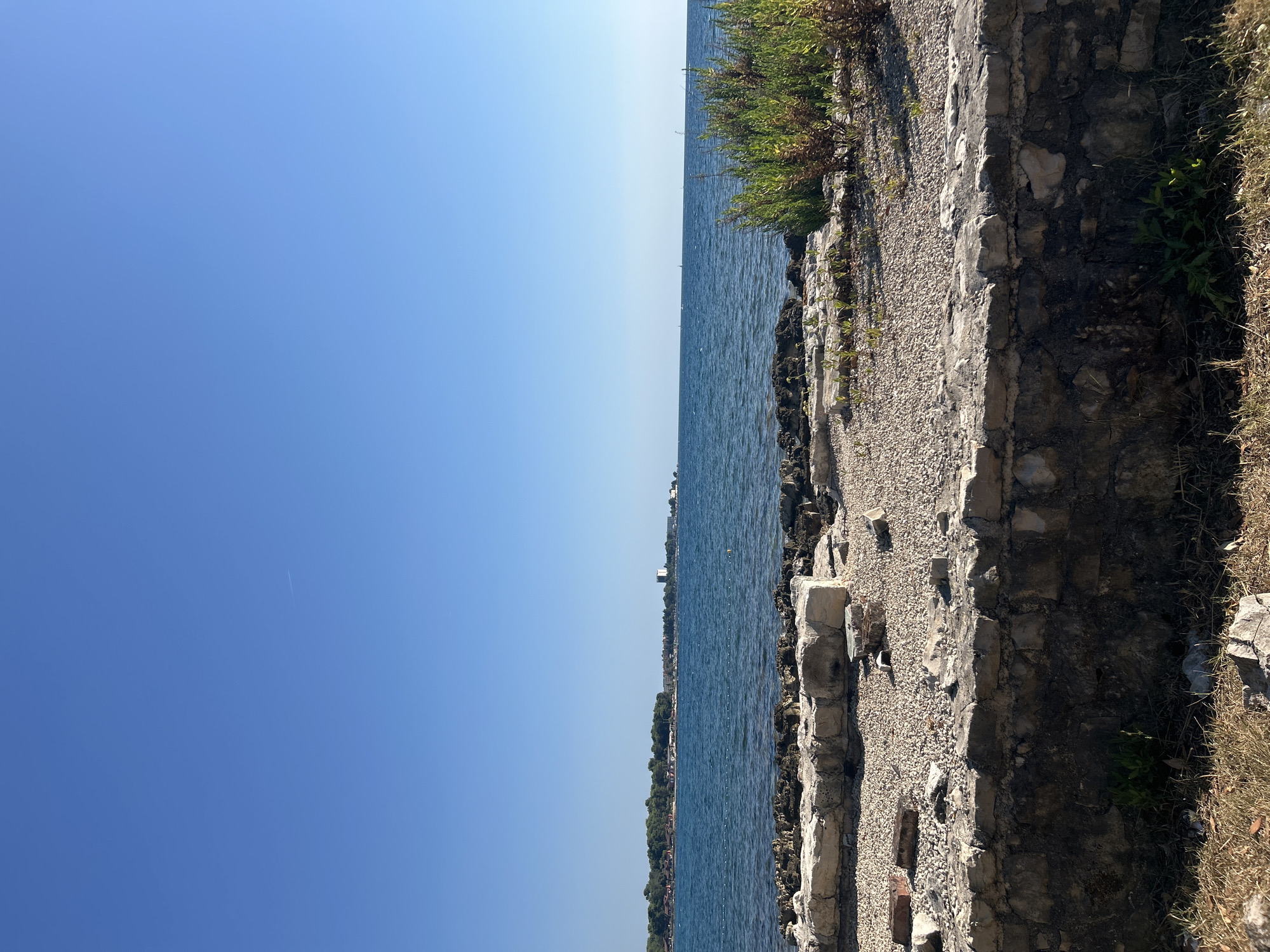 The image depicts a tranquil coastal scene with the clear blue sky dominating the upper half of the frame. Below the sky, a calm, deep blue sea stretches to the horizon. In the distance, a lush green shoreline is visible, dotted with buildings and trees. The foreground features a textured landscape of stones and gravel, bordered by a low, weathered stone wall. To the right, a cluster of green foliage adds a touch of natural vibrancy to the scene. The overall impression is one of a serene and picturesque seaside location in Katoro, Croatia.