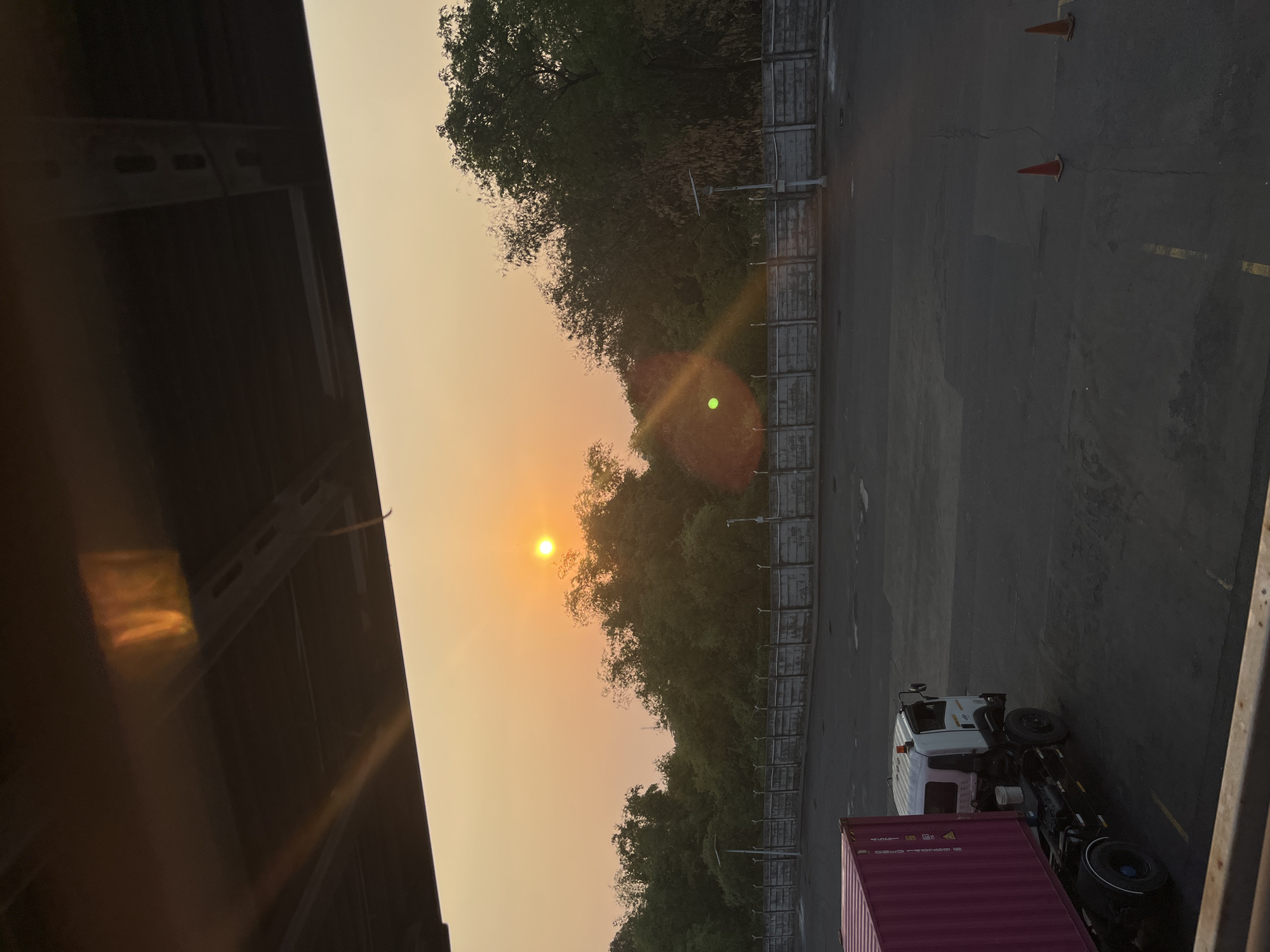 The image depicts an outdoor scene at what appears to be a logistics or industrial area. The time of day suggests either sunrise or sunset, with the sun positioned in the upper center of the frame, casting a warm, orange glow across the sky. In the foreground, a large, pink shipping container is partially visible on the left side, attached to a white truck with a black chassis. The truck has its headlights on, and a small orange light is visible on its roof.
Behind the truck, a grey concrete wall with a fence on top runs horizontally across the middle ground. Beyond the wall, a dense line of green trees and foliage forms a natural barrier. The ground in the foreground is dark asphalt, marked with faded yellow lines. Two orange traffic cones are placed on the asphalt in the right midground. Overhead, a dark, structural element, possibly part of an overpass or building, extends from the top edge of the image downwards, casting shadows and creating a framed view of the scene. The environment appears to be a functional, operational space within a city.
