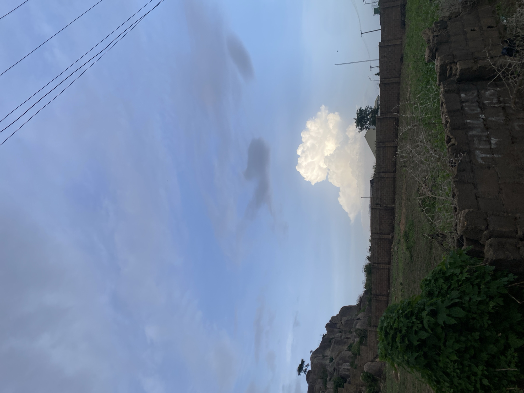 In the city of Barikin Jirga Summa, Nigeria, a natural landscape unfolds under a partly cloudy sky. In the foreground, a rough brick wall, partially constructed and bearing some graffiti, marks the edge of a plot of land. Beyond this, a dense cluster of green foliage dominates the left side of the frame, reaching upwards. Further back, a sturdy, brick wall stretches horizontally across the midground, delineating a property. Beyond this wall, a grassy expanse dotted with sparse vegetation extends towards a rocky outcrop on the left. This outcrop is steep and weathered, with a lone tree perched precariously near its summit. The sky above is a canvas of blues and whites, with several clouds scattered across it, including a prominent, fluffy white cumulus cloud that stands out against the lighter shades. Electrical wires crisscross the upper portion of the image, suggesting the presence of infrastructure. The scene appears to be an undeveloped or semi-developed area, characterized by natural rock formations and rudimentary construction.