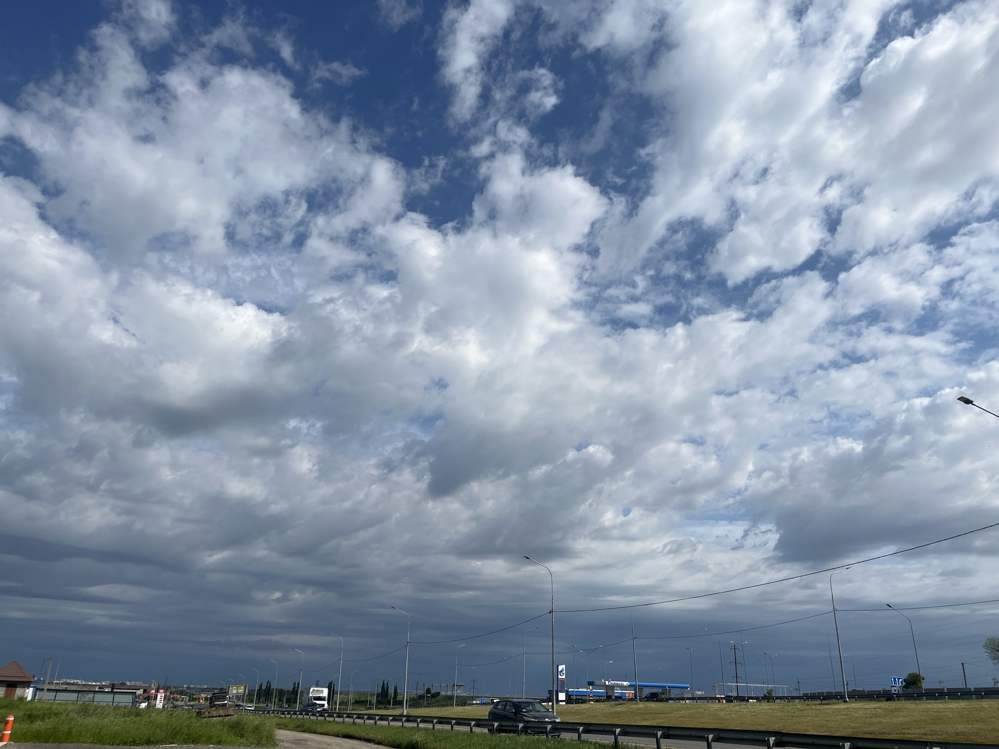 The image shows a partly cloudy sky over a highway in Adygeysk, Russia. The sky is mostly blue with large patches of white and gray clouds. The bottom third of the image is dominated by the highway, with a grassy median and guardrails. Streetlights line the road. Buildings and infrastructure are visible in the distance, including a gas station. A few vehicles are on the road, including a dark-colored car in the median. The overall scene depicts a typical roadside view in a suburban or semi-urban setting.