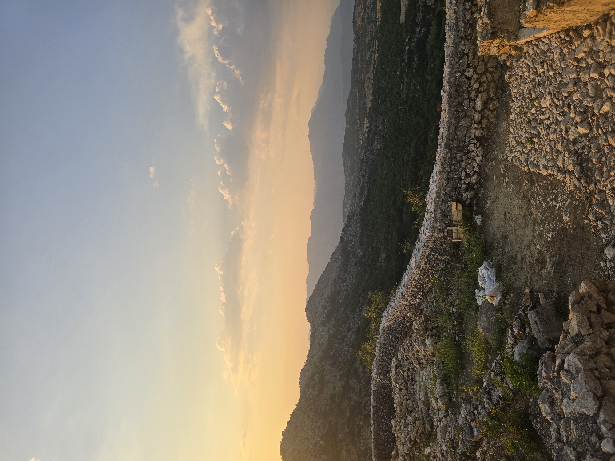 The image depicts a landscape view from a high vantage point, overlooking a valley with rolling hills and mountains in the distance. The foreground features a partially constructed or ruined stone wall, with loose rocks and vegetation growing around it. The ground is uneven and rocky, with patches of dry soil and grass. In the midground, a dense forest covers the slopes of the hills, giving way to more arid, rocky terrain further up. The sky is a gradient of soft oranges and yellows near the horizon, transitioning to a pale blue overhead, with scattered clouds. The scene is bathed in the warm light of what appears to be a sunset or sunrise. The location is identified as Gornja Vrbica, Montenegro. There are no people or animals visible in the image. The overall impression is one of a rugged, natural environment with remnants of human construction.