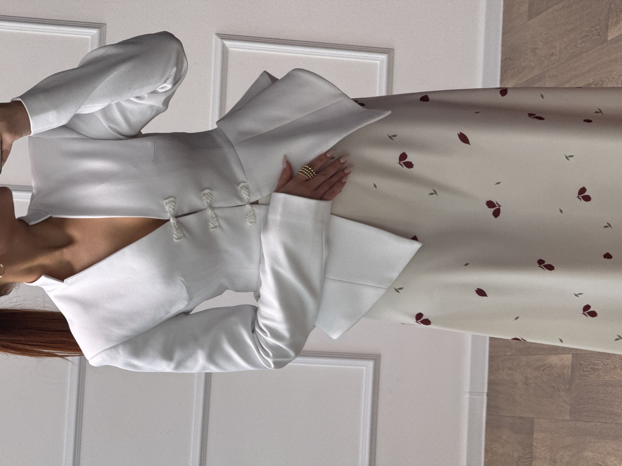 An indoor scene in Chaikovsky, Russia, depicts a partially visible individual from the mid-torso to mid-calf, captured in a mirror selfie. The person, gender unidentified but with visible reddish-brown hair tied in a ponytail, wears a white, V-neck, long-sleeved jacket featuring a mandarin collar and three decorative white frog closures down the front. The jacket's lower portion flares out in a peplum style. Beneath the jacket, a light cream-colored skirt with a scattered pattern of small dark red leaf-like shapes and tiny dark gray 'T'-shaped motifs is visible. The individual's right hand is raised, holding a smartphone (partially obscured) to take the photograph, while the left hand rests at the side, displaying a multi-band gold-colored ring on the ring finger. The fingernails are manicured with a light pink polish. Small gold-colored hoop earrings are visible on the right ear. The background consists of a plain white wall adorned with rectangular white decorative molding. The floor is made of light brown wooden planks.