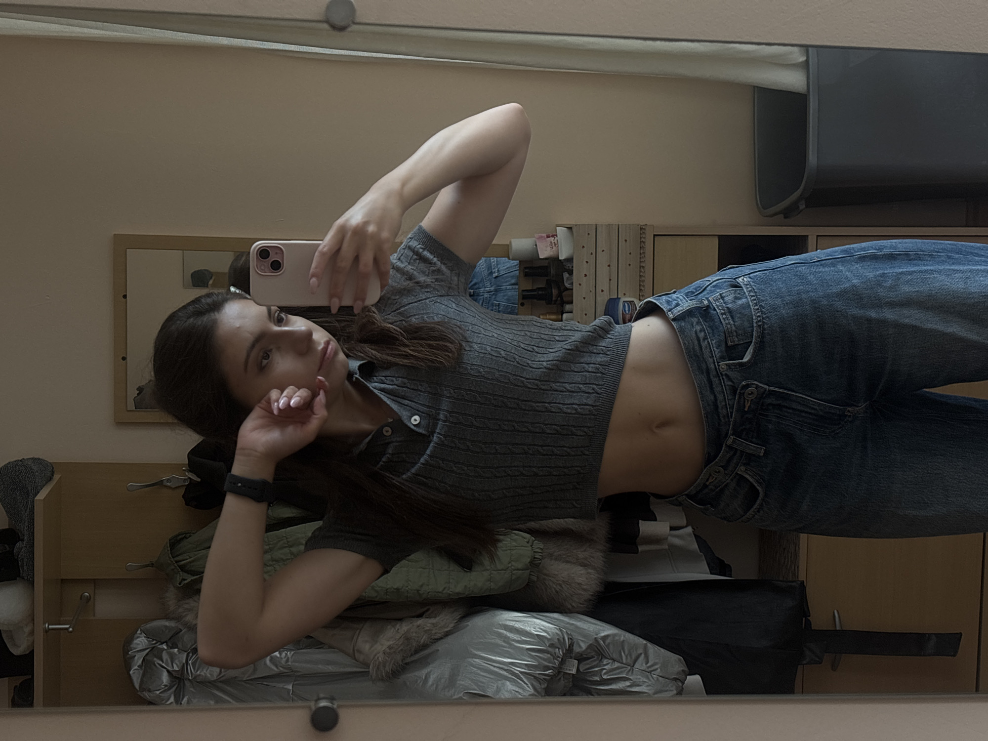 A young woman with long, dark hair is lying down, taking a selfie in a mirror. She is wearing a gray, short-sleeved, cable-knit cropped shirt and blue denim jeans. Her left arm is bent, supporting her head, while her right hand holds a pink smartphone. The woman's midriff is exposed, and she is wearing a dark-colored fitness tracker on her left wrist.
The setting appears to be a bedroom or dressing area. In the background, reflected in the mirror, are shelves with various items, including what appear to be stacked clothes or bags, and a framed picture. The lighting suggests it is daytime. The overall mood is casual and relaxed. There is no visible text in the image. The location is Varna, Bulgaria.