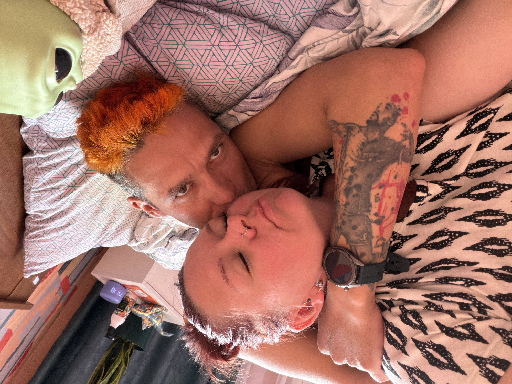 Two individuals are lying in bed, one appearing to kiss the other on the cheek. The individual kissing has orange hair and is looking at the camera. The other individual has red hair and their eyes are closed. The individual on the bottom is wearing a black smartwatch on their left wrist and has a tattoo on their left arm. An alien mask is visible in the upper left corner of the image. A digital clock on a nightstand reads "14:18." The location appears to be a bedroom, and the image does not offer information about being located in the city of Chaikovsky, Russia.