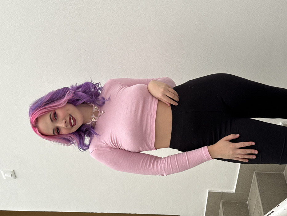 A female individual is depicted standing against a plain, light-colored wall, positioned slightly right of the frame's center, within what appears to be an indoor stairwell setting in the City of Zagreb, Croatia. She has wavy, shoulder-length hair, dyed vibrant pink on the upper layers and purple on the lower layers. Her facial expression is a slight smile, revealing teeth. She wears dark red lipstick and visible eye makeup. Her attire includes a long-sleeved pink crop top that exposes her midriff, paired with black fitted pants. A translucent choker necklace adorned with white circular beads and a small silver-colored heart pendant is worn around her neck. Her left hand rests on her left hip, while her right arm is partially out of view. A grey staircase with a white railing is visible in the lower left background, and a white rectangular object, possibly a light switch, is mounted on the wall in the upper left.