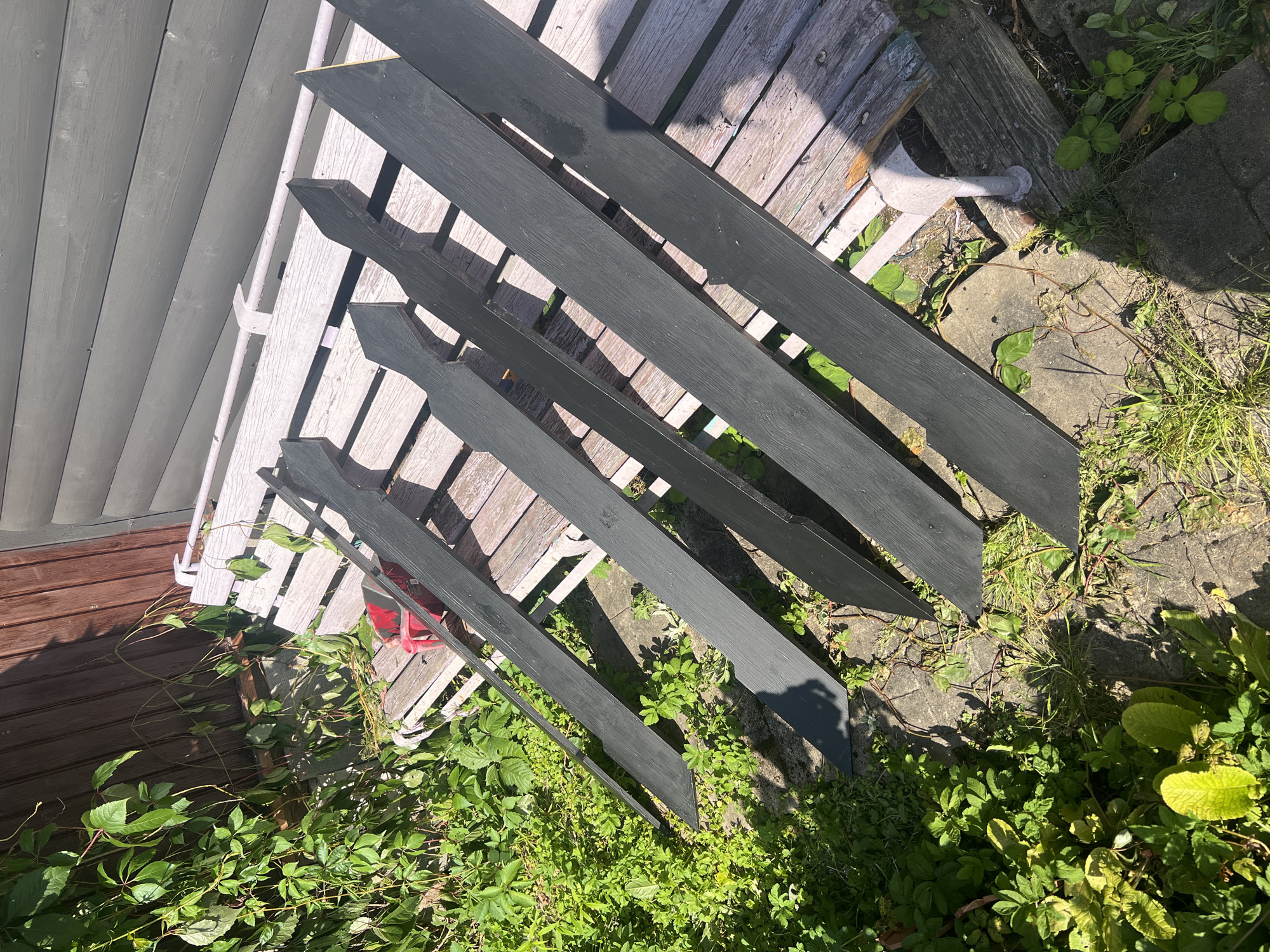 A high-angle, eye-level medium shot captures a garden scene with several newly painted black wooden planks laid out on a weathered wooden bench. The planks are pointed at one end, resembling a picket fence or decorative garden border. They are arranged in a fanned-out pattern across the bench. To the left of the bench, lush green foliage and vines grow, some of which are draped over the edge of the bench and onto the planks. Behind the bench is a brown wooden fence or wall, which appears to be a side of a building. The ground in front of the bench is a mix of concrete pavers and dirt, with more greenery sprouting through the cracks and around the edges. The scene is brightly lit by sunlight, casting shadows from the planks and foliage. The location is identified as Rosа Snt, Russia.