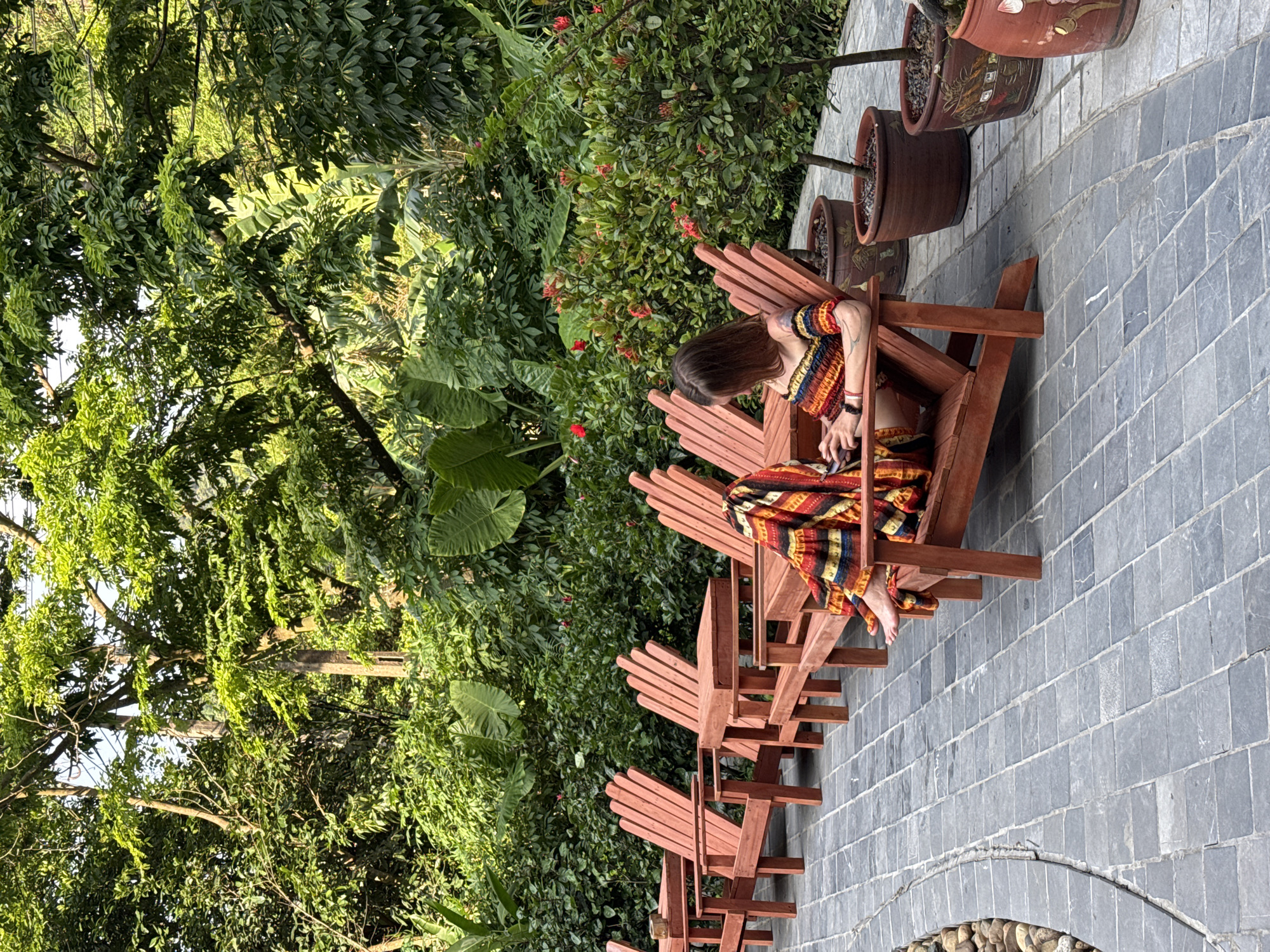In Nam Hoa Lư Ward, Vietnam, a woman is seated in a wooden chair on a stone patio. She is wearing a colorful, patterned outfit and is looking down at her phone. The setting appears to be a lush garden with abundant greenery and plants, including large-leafed tropical plants and flowering bushes with red blooms. Several other empty wooden chairs are arranged in a row behind her. Large ceramic pots containing plants are visible on the patio to the right of the woman. The scene is illuminated by natural light, suggesting it is daytime. The atmosphere seems peaceful and relaxed.