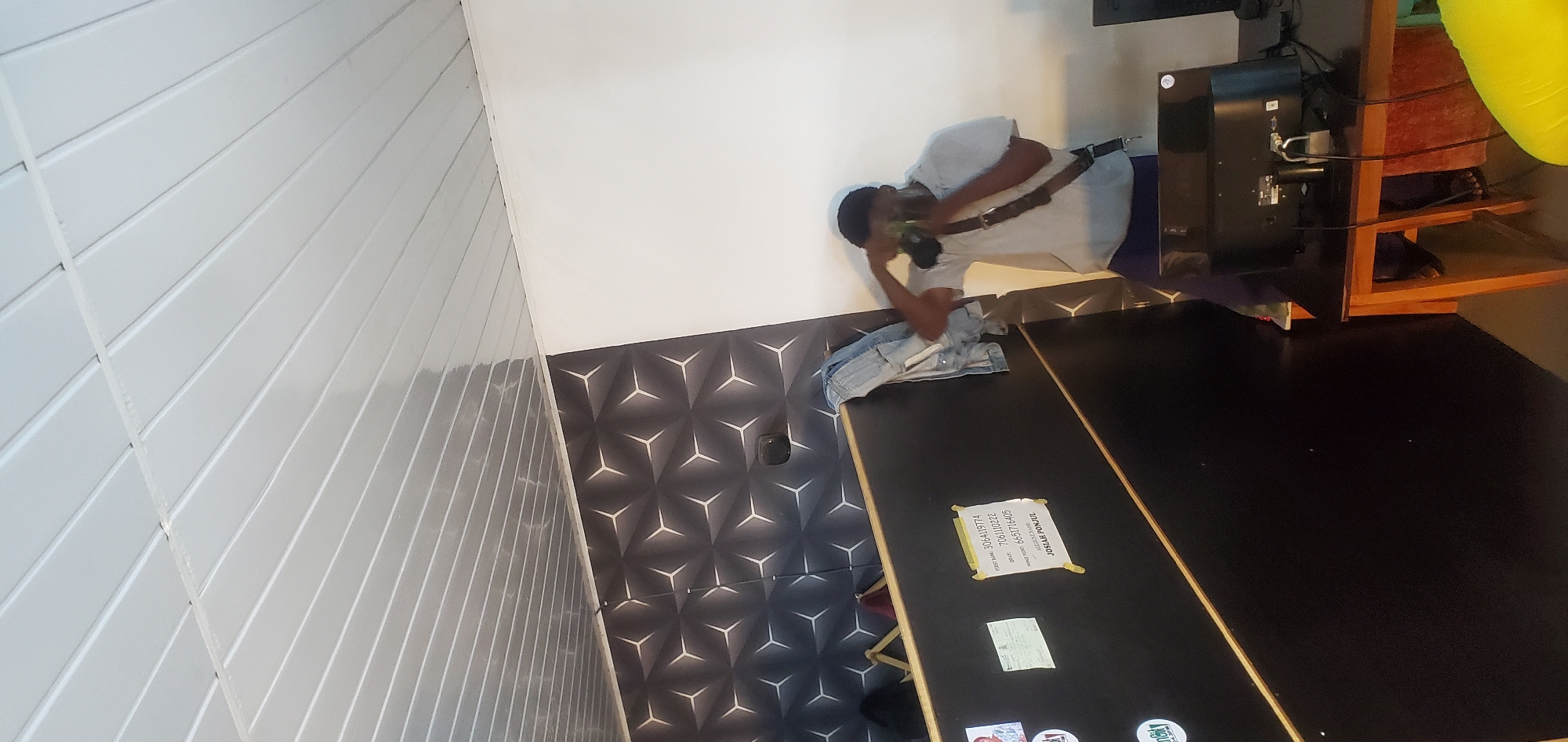In Bukuru, Nigeria, a person wearing a light grey t-shirt, dark blue pants, and a white cap is holding a green object to their face, appearing to be taking a picture. They are standing in front of a black counter with a patterned wallpaper behind it. A denim jacket is draped over the counter. A computer monitor is visible on a wooden desk to the right, with cables connected to it. The environment includes a white wall and a textured ceiling. Several papers with writing are attached to the counter.
