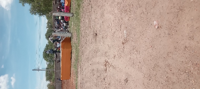 The image shows a dirt field in Kitengela, Kenya. A large, orange, rectangular structure is visible in the background, partially obscured by a low concrete wall. There are a number of people standing on a platform in front of the structure, including a group of men in suits and a group of women in bright, colorful dresses. The ground is dusty and there are several sets of footprints visible in the dirt. The sky is blue with a few white clouds. The location is a large open space with a few trees in the background.