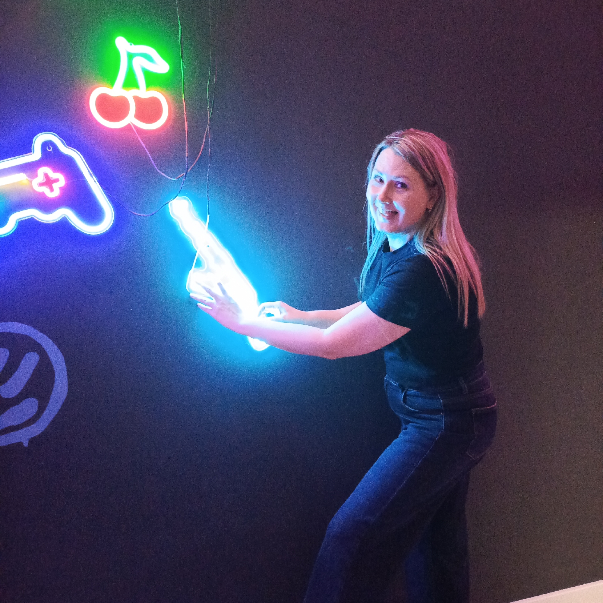A woman stands in front of a dark wall adorned with neon signs. The signs, from left to right, include a graffiti-style "W", a game controller, cherries, and a hand pointing upwards. The woman is fair-skinned with blonde hair streaked with pink. She wears a black t-shirt and jeans and appears to be holding the neon hand sign. The scene takes place indoors, but the exact location in Chaikovsky, Russia, cannot be determined from the image.