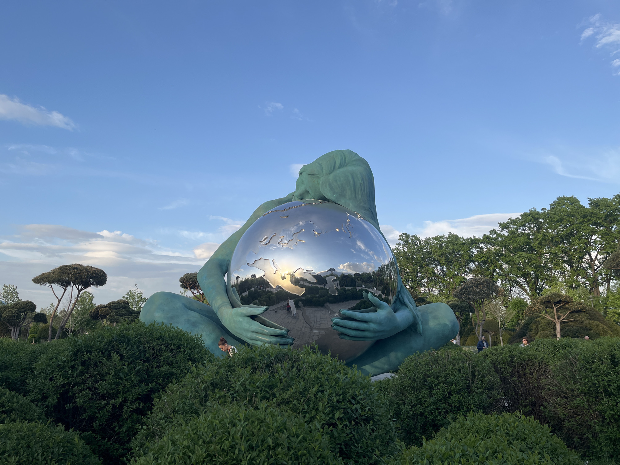 A full, eye-level shot depicts a large, turquoise-colored sculpture of a seated woman embracing a reflective, spherical object, likely a globe. The sculpture is situated amidst lush, green shrubbery in a park-like setting, with trees visible in the background. The sky above is blue with scattered clouds. The sculpture's reflective surface mirrors the surroundings, including trees and buildings. In the background, there are a few pedestrians. The scene appears to be in Krasnodar, Russia, given the location of the sculpture.