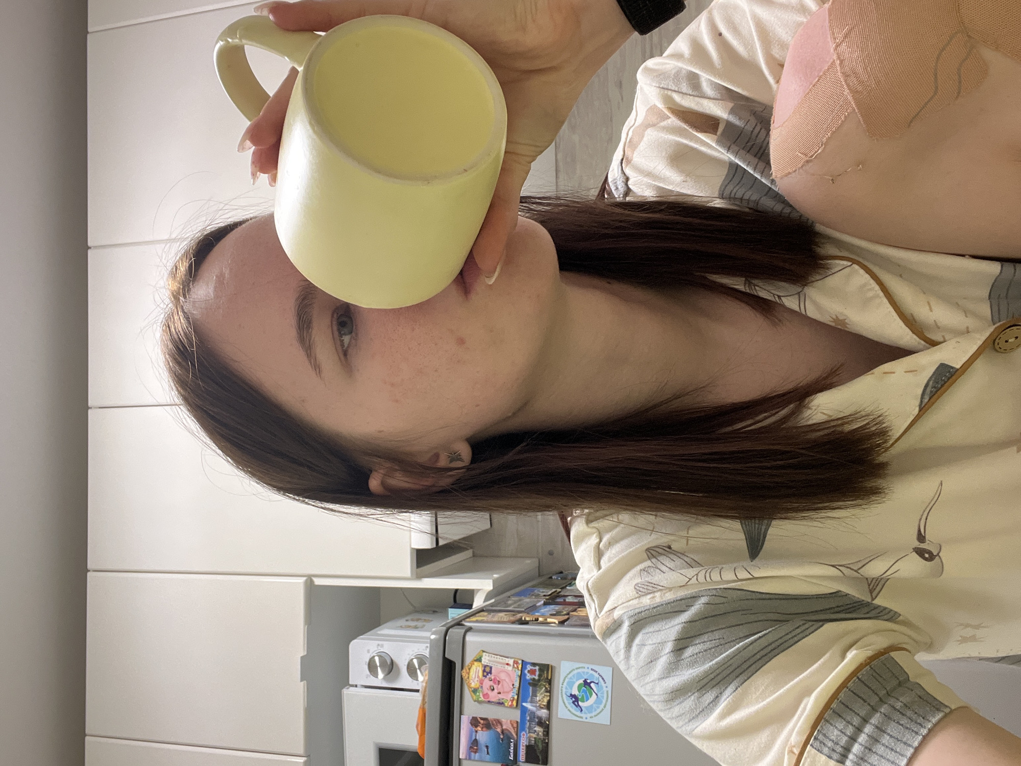 A young woman is captured in a close-up, sideways-oriented shot, appearing to be in a kitchen in Chaikovsky, Russia. She has long, dark brown hair and fair skin with visible freckles. She is dressed in light-colored pajamas adorned with a patterned print featuring abstract shapes and what appear to be birds or fish. A star-shaped earring is visible in her left ear.
The woman holds a light yellow ceramic mug to her lips with her right hand, suggesting she is in the process of drinking from it. Her fingernails are long and manicured. On her upper right arm or shoulder, several strips of light-colored medical tape or kinesiology tape are visible, indicating a possible injury or recovery.
In the background, a modern kitchen setting is discernible, featuring white, handle-less cabinets and a silver-toned refrigerator. The refrigerator is decorated with several magnets, including one featuring a globe with two figures, and another partially displaying the word "Gold" along with some Russian text. Part of a white microwave oven is also visible next to the refrigerator. The overall scene suggests a casual, domestic moment, likely in the morning or evening given her attire.