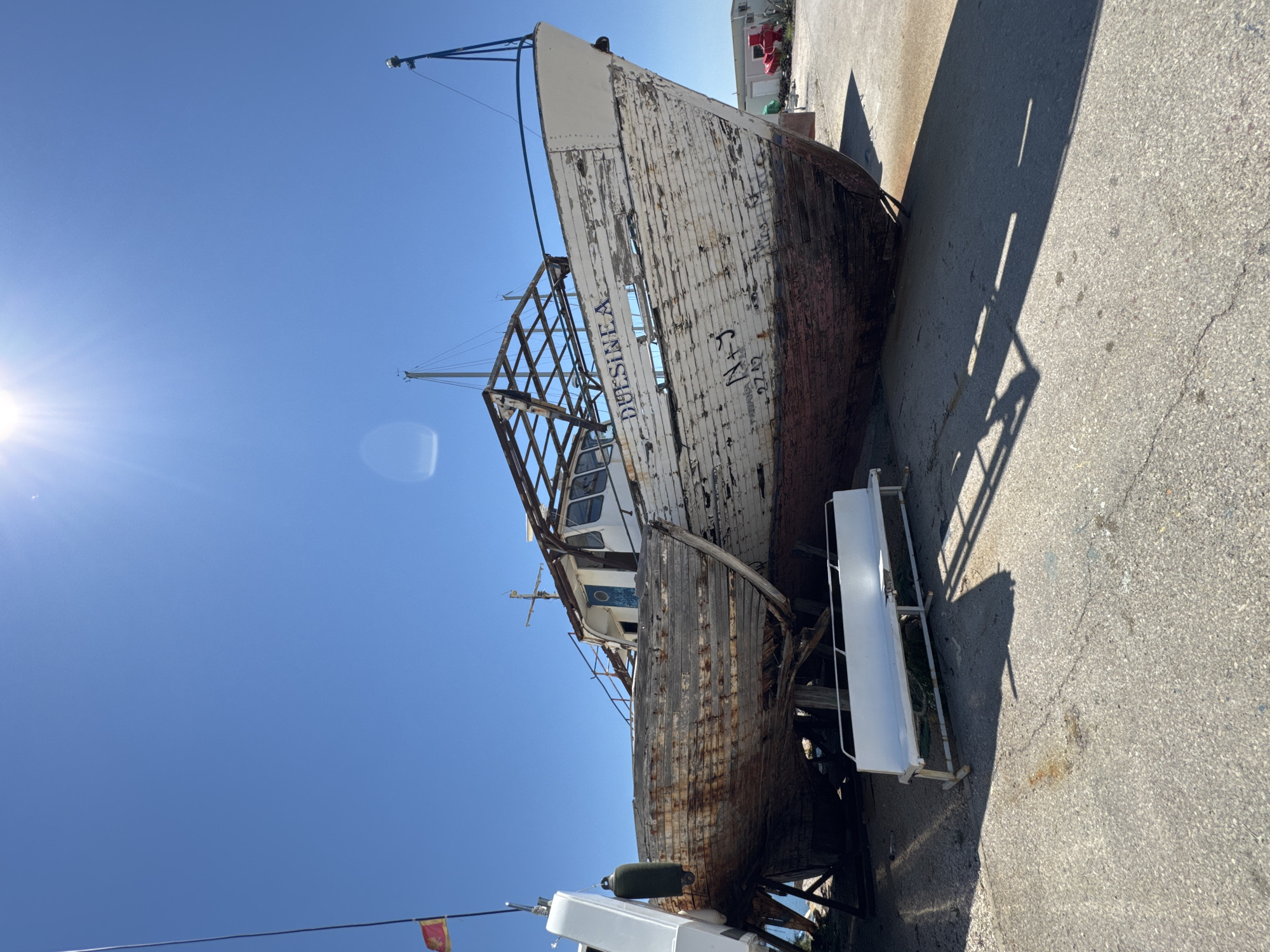 The image depicts an abandoned wooden boat undergoing apparent restoration or repair, positioned on dry land under a clear, sunny sky. The boat, weathered and showing signs of disrepair, has a partially dismantled superstructure. The name "DUESINEA" is visible on the side of the hull, along with other markings. The boat rests on wooden supports. In the foreground, a white metal bench is visible, casting a shadow on the concrete ground. In the background, there are structures that appear to be sheds or workshops, and some red equipment, possibly related to the boatyard. A red and yellow flag is attached to a pole on the left side of the image. The overall environment suggests an outdoor boatyard or shipyard. The scene is devoid of any visible people. The context of Bar, Montenegro is not directly evident from the visual information alone but would be inferred from external knowledge.
