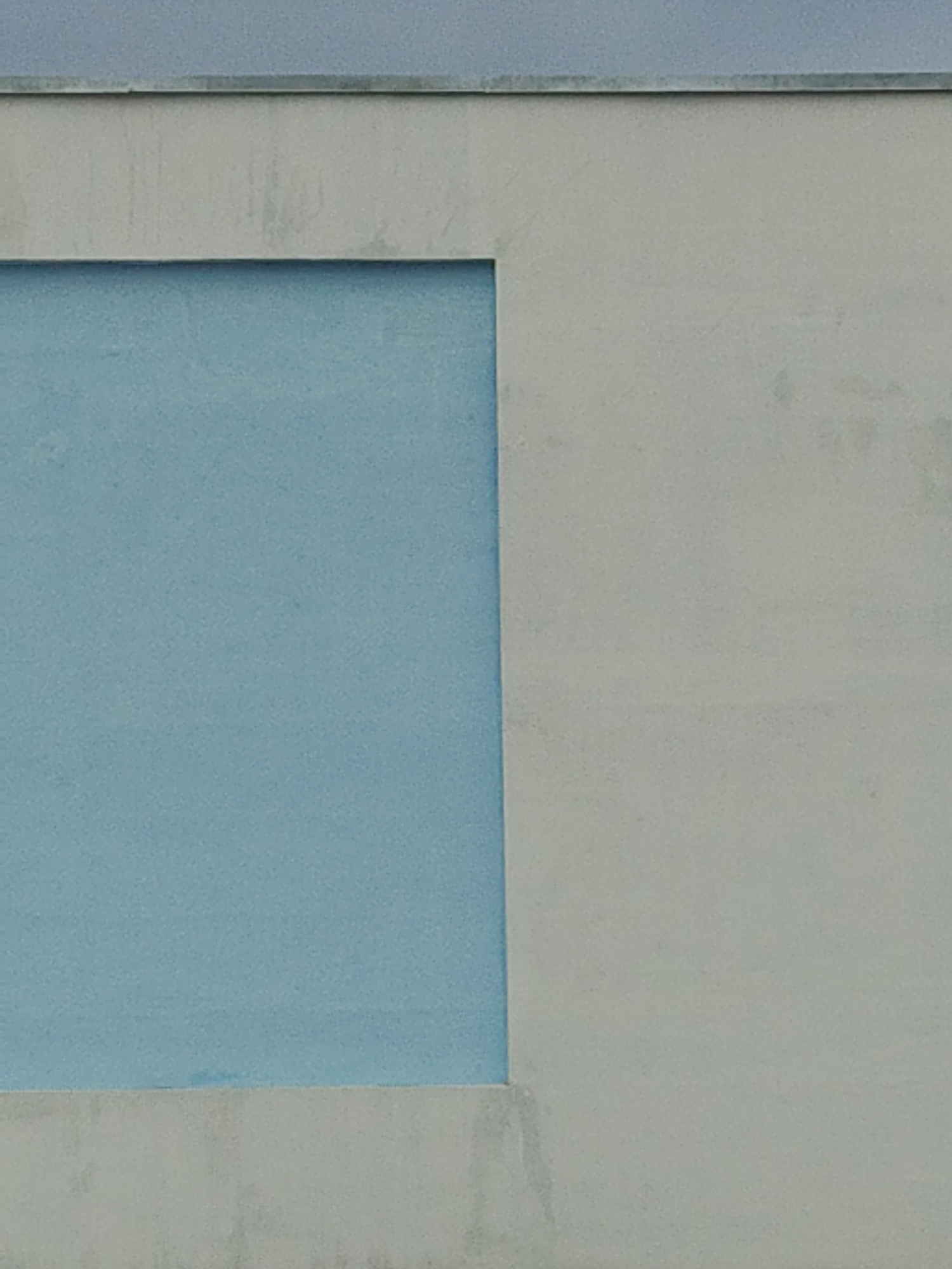 A close-up shot shows a wall with a rectangular recess filled with a light blue material. The wall itself is a pale, off-white color with a slightly rough texture. A horizontal metal strip runs along the top edge of the wall. The wall appears to be an exterior wall, possibly part of a building. The location is unknown.
