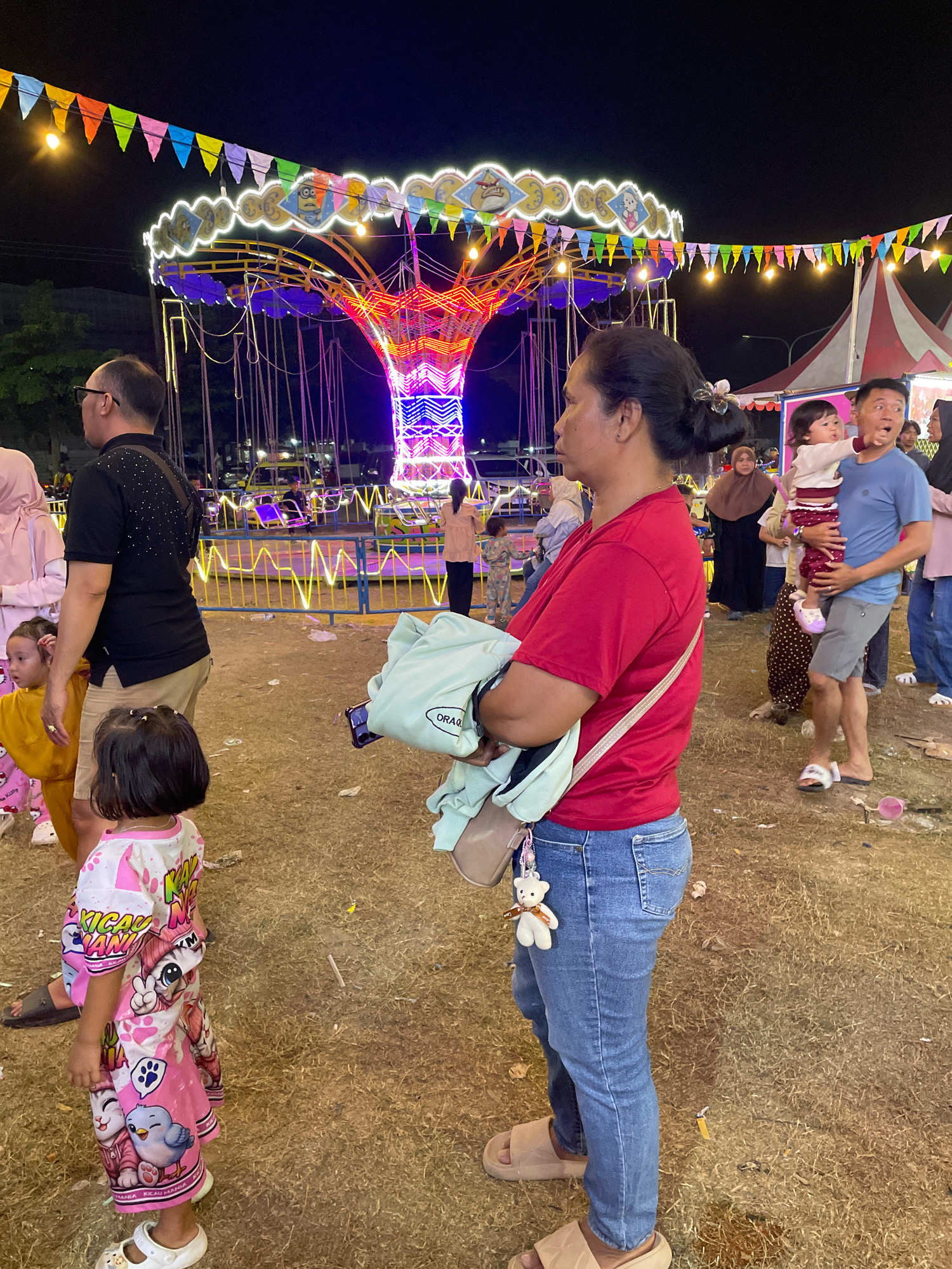An outdoor night scene at an amusement park or fairground in Pulung Kencana, Indonesia, features a vibrant carousel with colorful lights in the background. Several people are visible, including a woman in a red shirt and blue jeans carrying a light blue bag, and a man in a blue shirt and grey shorts holding a young child. Other individuals and children are scattered throughout the frame, some appearing to be watching the rides or moving around the grounds. The area is covered in dry grass, and scattered debris is visible on the ground, suggesting a well-used public space. Decorative banners and string lights adorn the fairground, adding to the festive atmosphere.