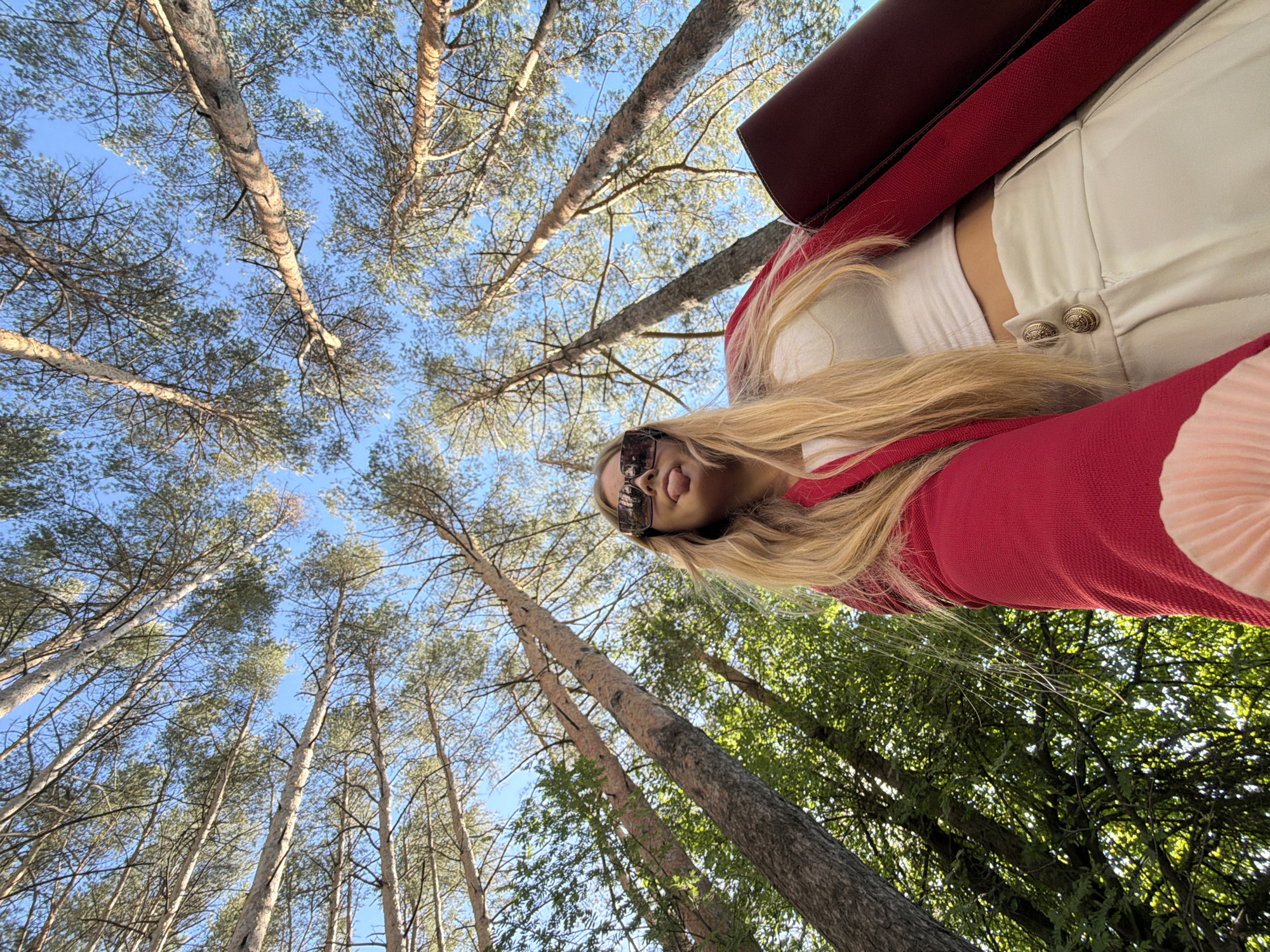 In Izhevsk, Russia, a low-angle shot captures a young woman with long blonde hair, wearing large sunglasses, a white crop top, a red jacket, and white pants, with her tongue playfully sticking out. She is holding a burgundy folder. The background is dominated by tall pine trees reaching towards a clear blue sky. Sunlight filters through the sparse branches, creating a dappled effect on the forest floor. The image is taken from a perspective looking up at the woman and the trees, emphasizing their height and the expansive sky.