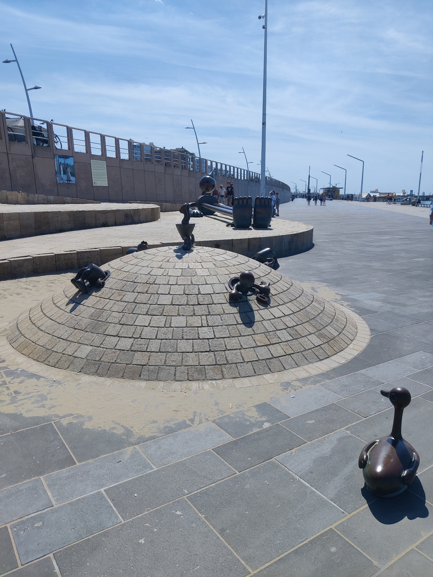The image depicts a sunny day at the Scheveningen boulevard in The Hague, Netherlands. In the foreground, a bronze sculpture of a duck stands on a paved walkway made of large grey stones. Behind the duck sculpture, a rounded, brick-like structure serves as a base for several bronze sculptures of human figures in various contorted positions. These figures appear to be interacting with or climbing on the structure. The background reveals a pedestrian promenade with people walking, a raised walkway with railings, lampposts, and buildings further in the distance. The sky is blue with scattered clouds.