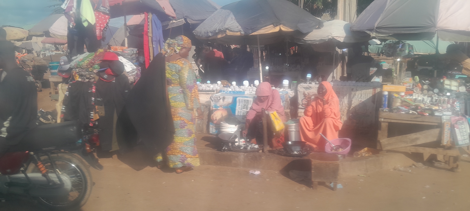 In Nyanya, Nigeria, a bustling marketplace scene unfolds under a clear sky, with a focus on two women selling food. The woman on the left, wearing a pink hijab and a patterned dress, is bent over a cooking pot, appearing to be engaged in preparing food. Beside her, another woman in a vibrant orange hijab and dress sits, observing the activity or interacting with customers. The area is lined with market stalls covered by umbrellas, displaying various goods like clothing and what appear to be electronics or household items. A motorcycle is parked on the left side of the frame, and the ground is a dusty, reddish-brown earth. The overall atmosphere is one of everyday commerce and activity within the Nigerian city.